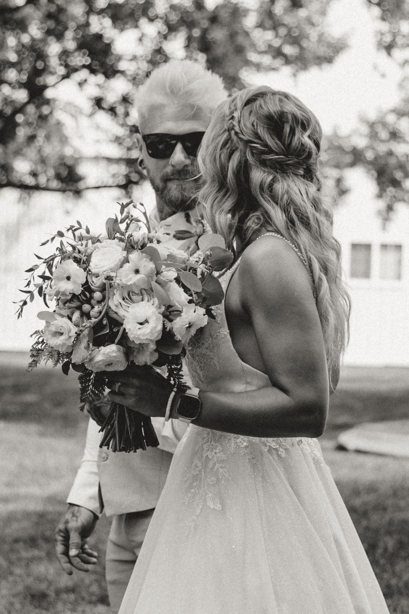 Black and white photo of a bearded groom in sunglasses holding a floral bouquet beside his bride in a lace wedding gown.