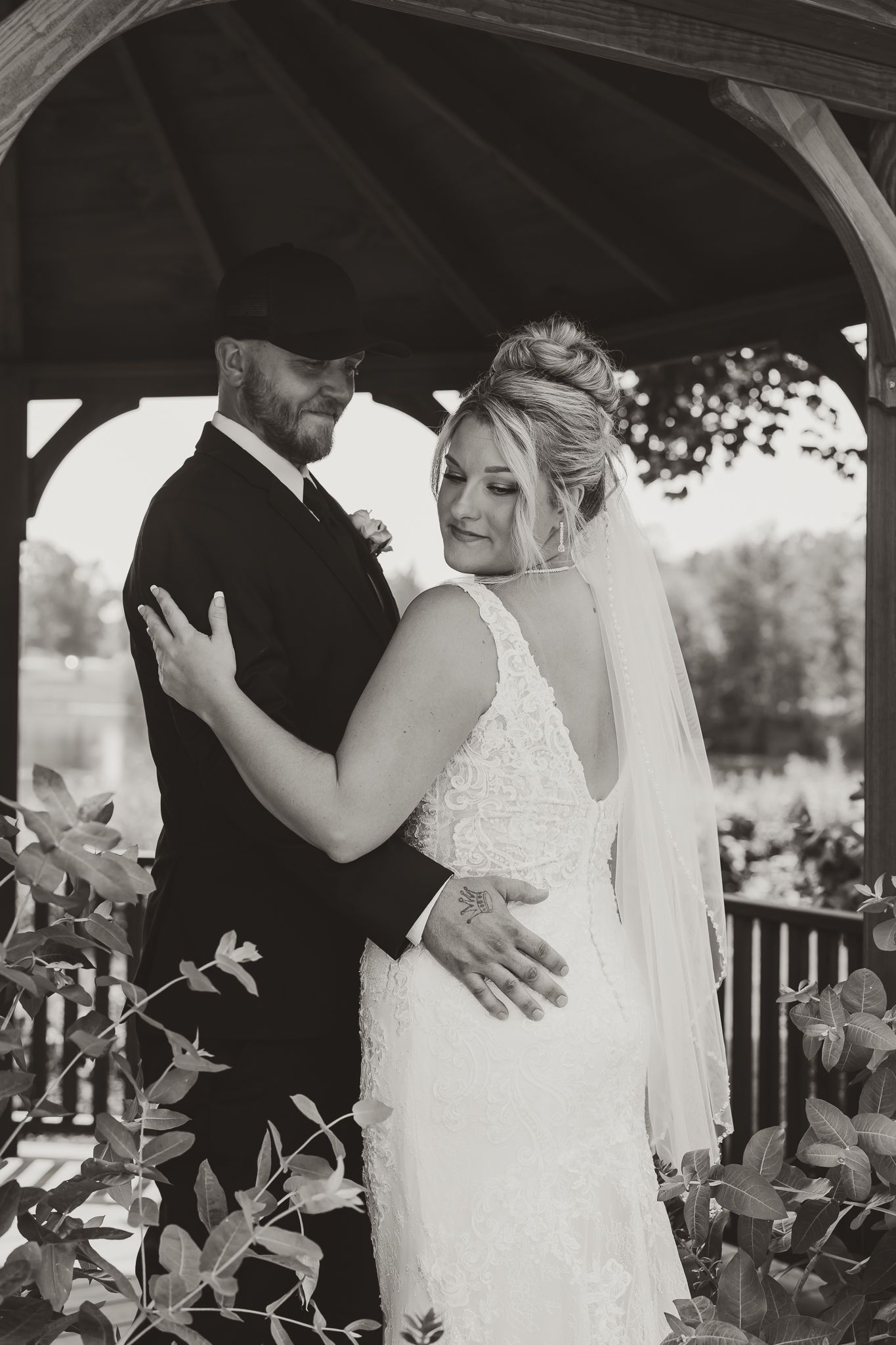 Black and white photo of a bride and groom embracing under a wooden gazebo, surrounded by lush greenery.