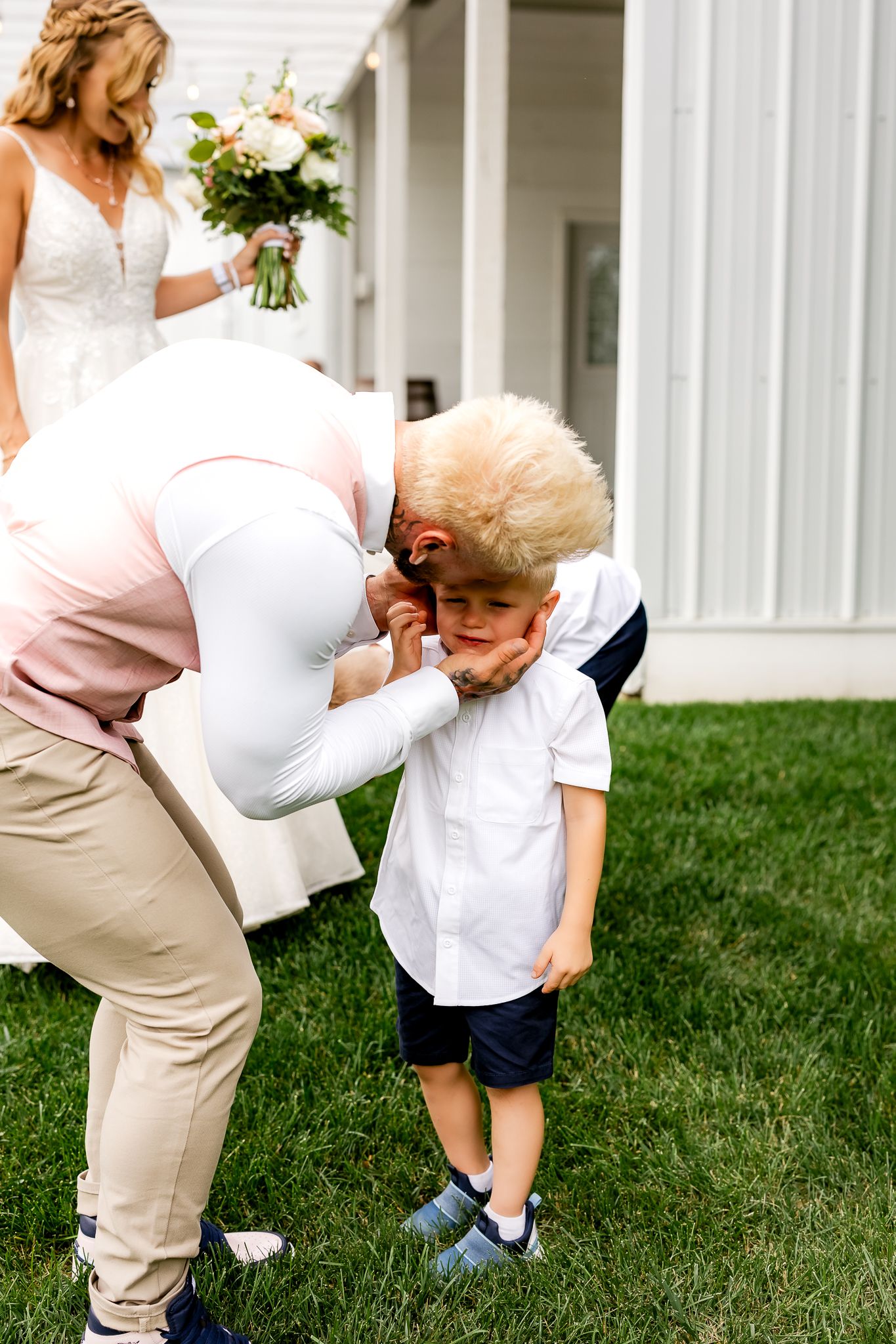 Adult in pink jacket adjusts young blonde boys collar at outdoor wedding ceremony on green grass.