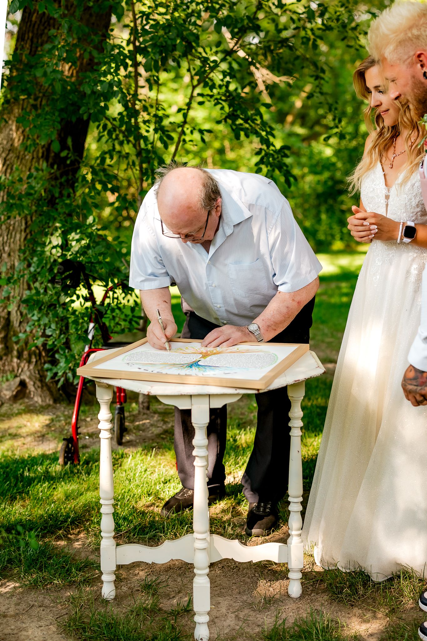 Elderly man signing wedding documents at a white table outdoors, beside a bride in a white dress.