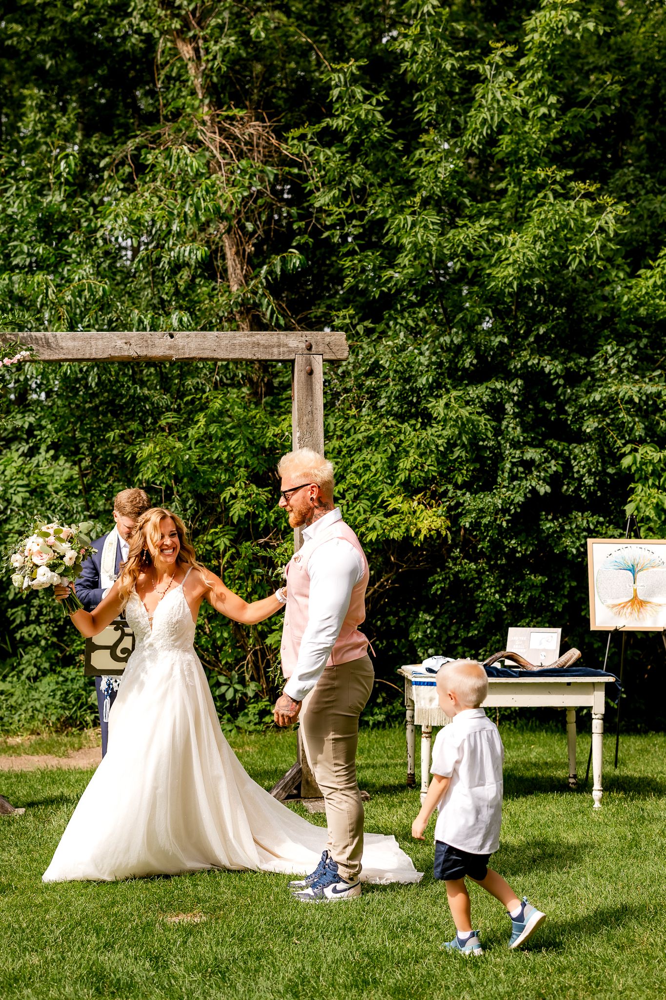 Bride and groom holding hands during outdoor wedding ceremony on green lawn with young child nearby.