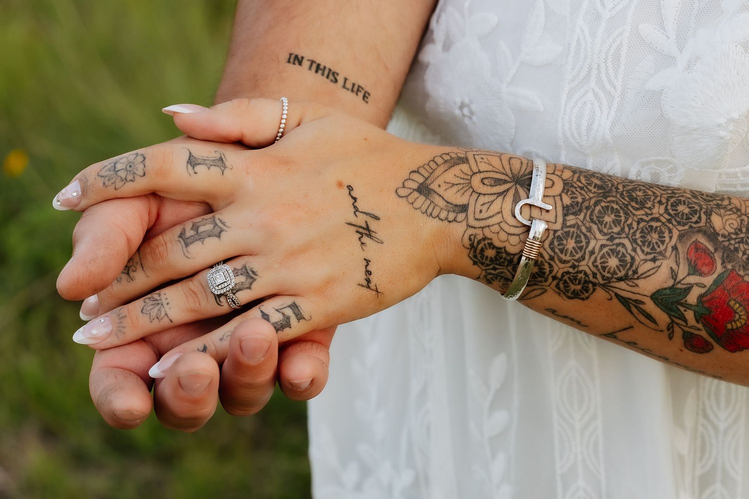 Close-up of couples intertwined tattooed hands, woman wearing white lace dress and delicate rings.
