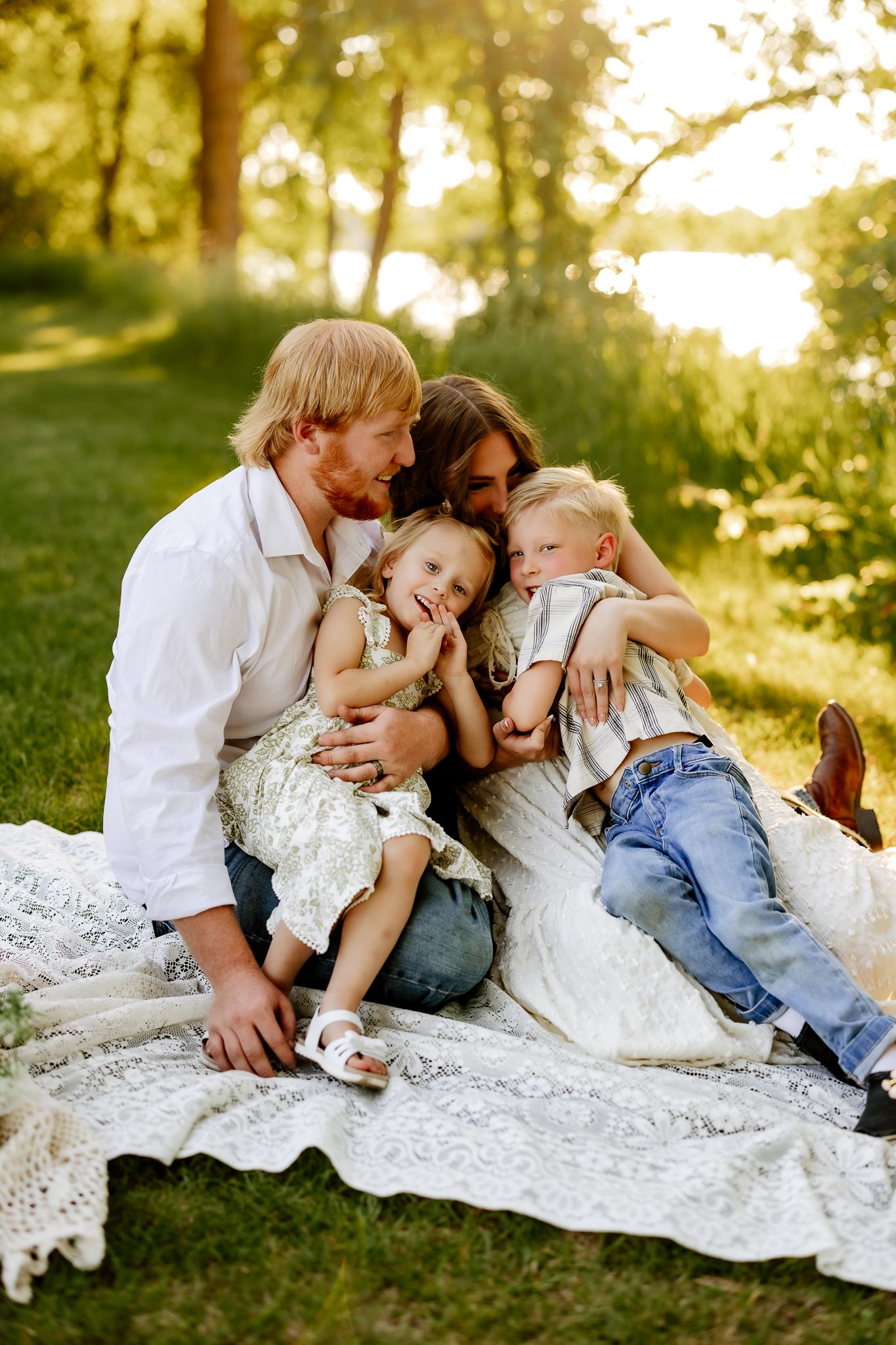 A loving family of four cuddles together on a lace blanket in a sunny green park near a lake.