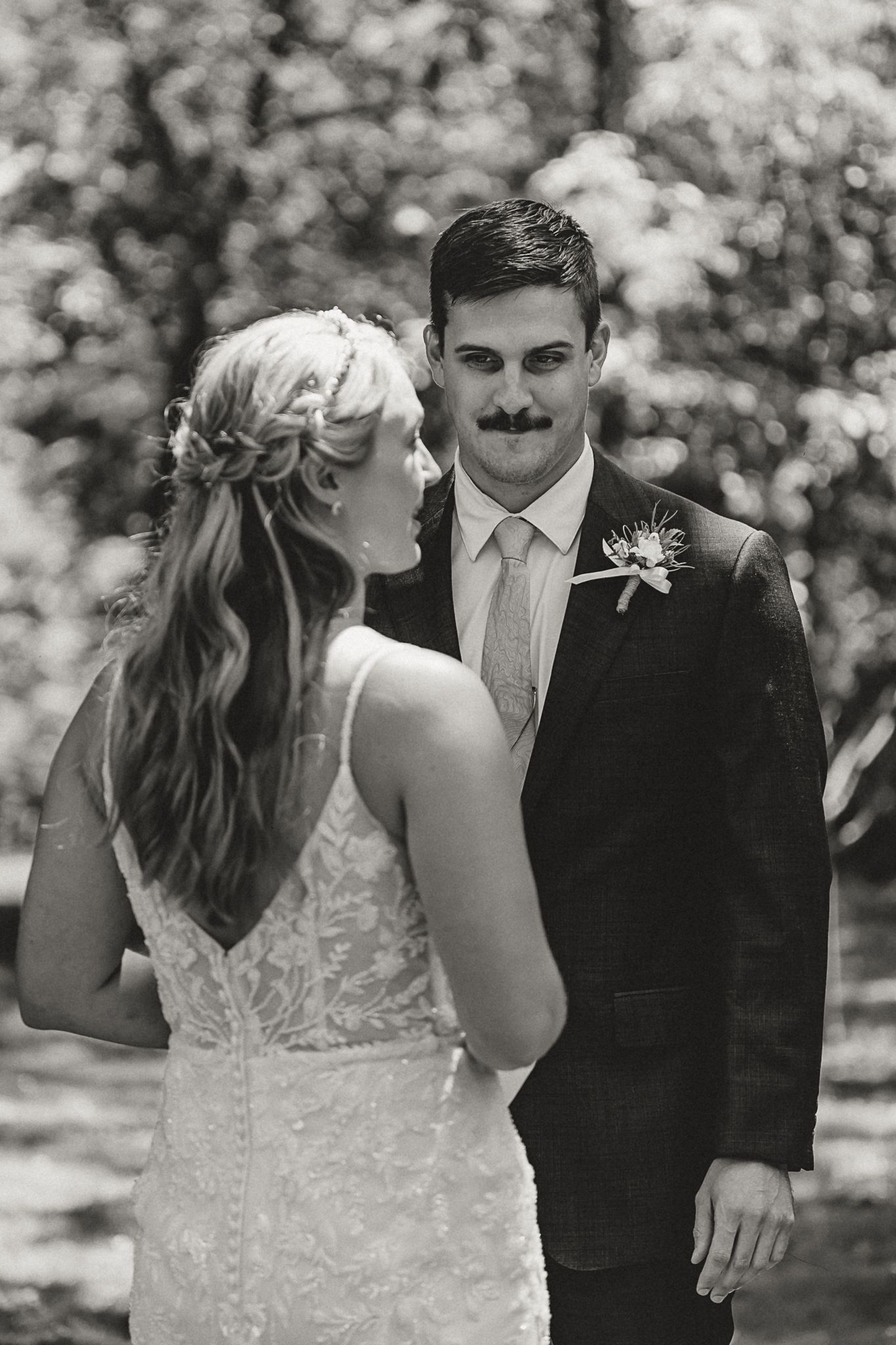 Black and white portrait of bride and groom sharing a tender first look moment among trees outdoors.