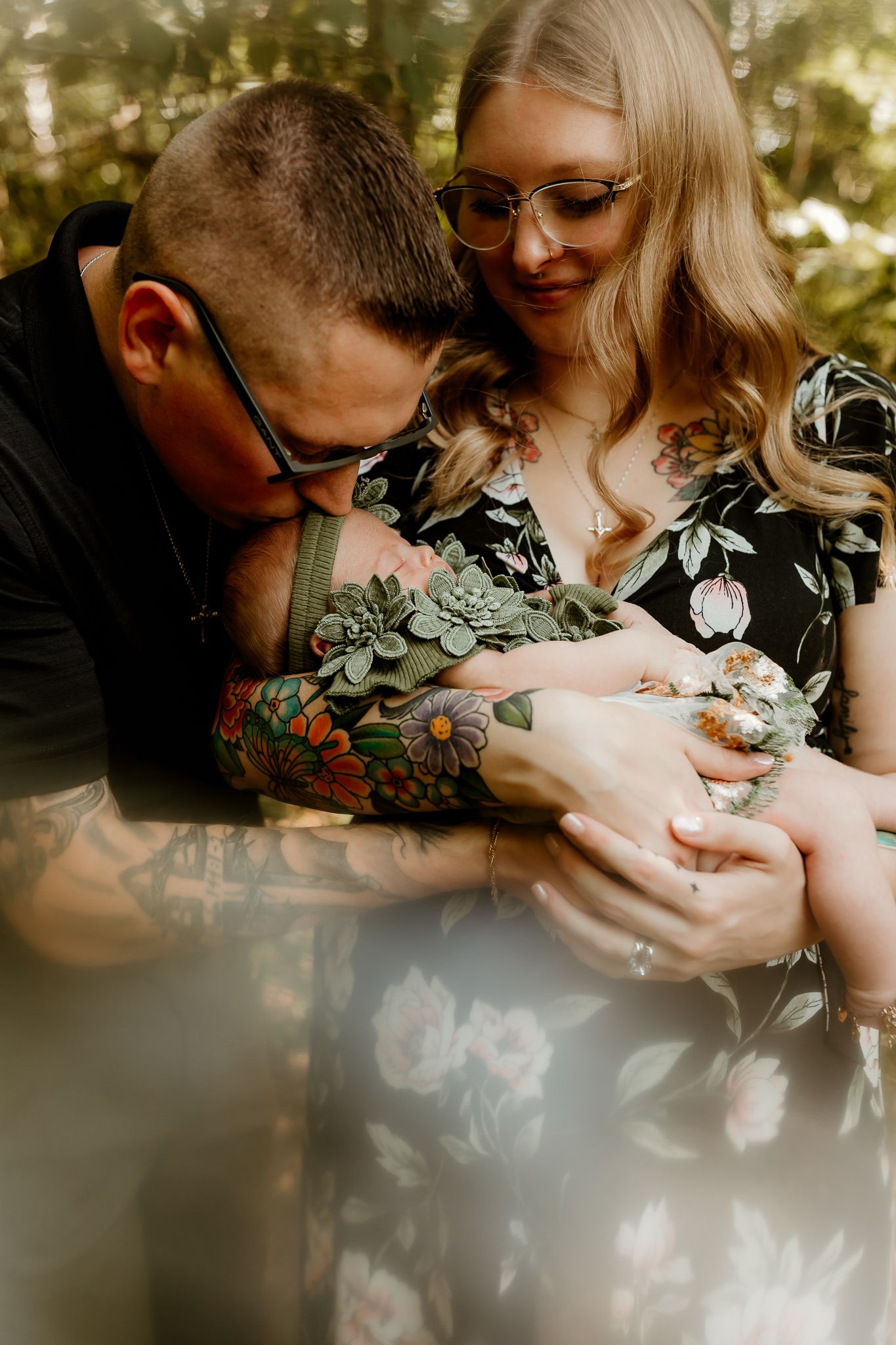 Couple with tattoos tenderly holding newborn baby outdoors, man kissing infant while woman in floral dress smiles softly.