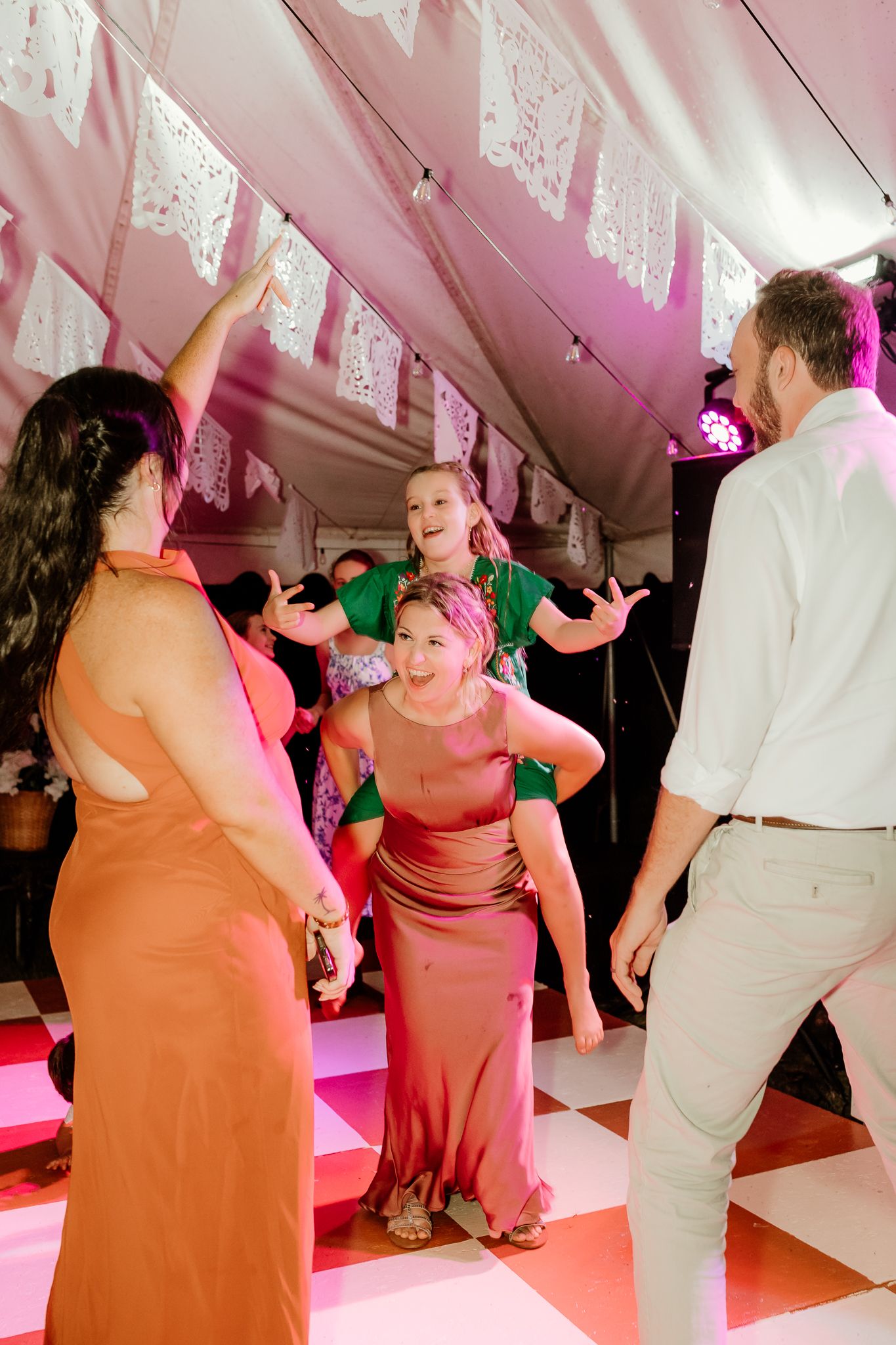 Guests dancing joyfully at a wedding reception under decorative papel picado banners in a pink-lit tent.