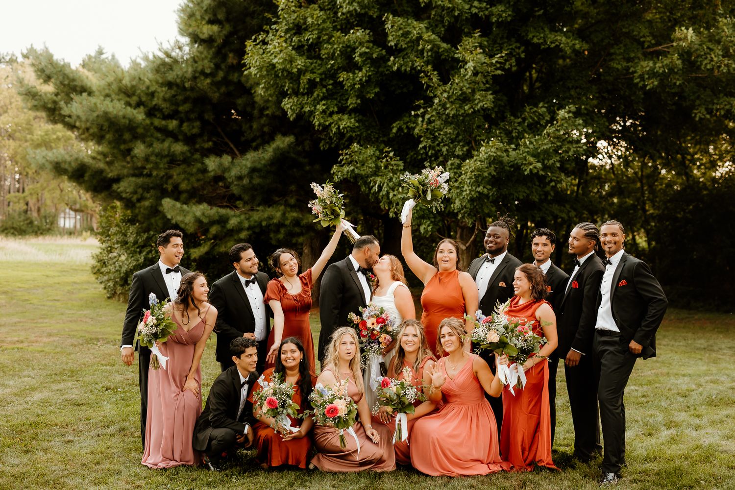 Wedding party in formal attire poses outdoors on green lawn, bridesmaids in coral dresses, groomsmen in black tuxedos.