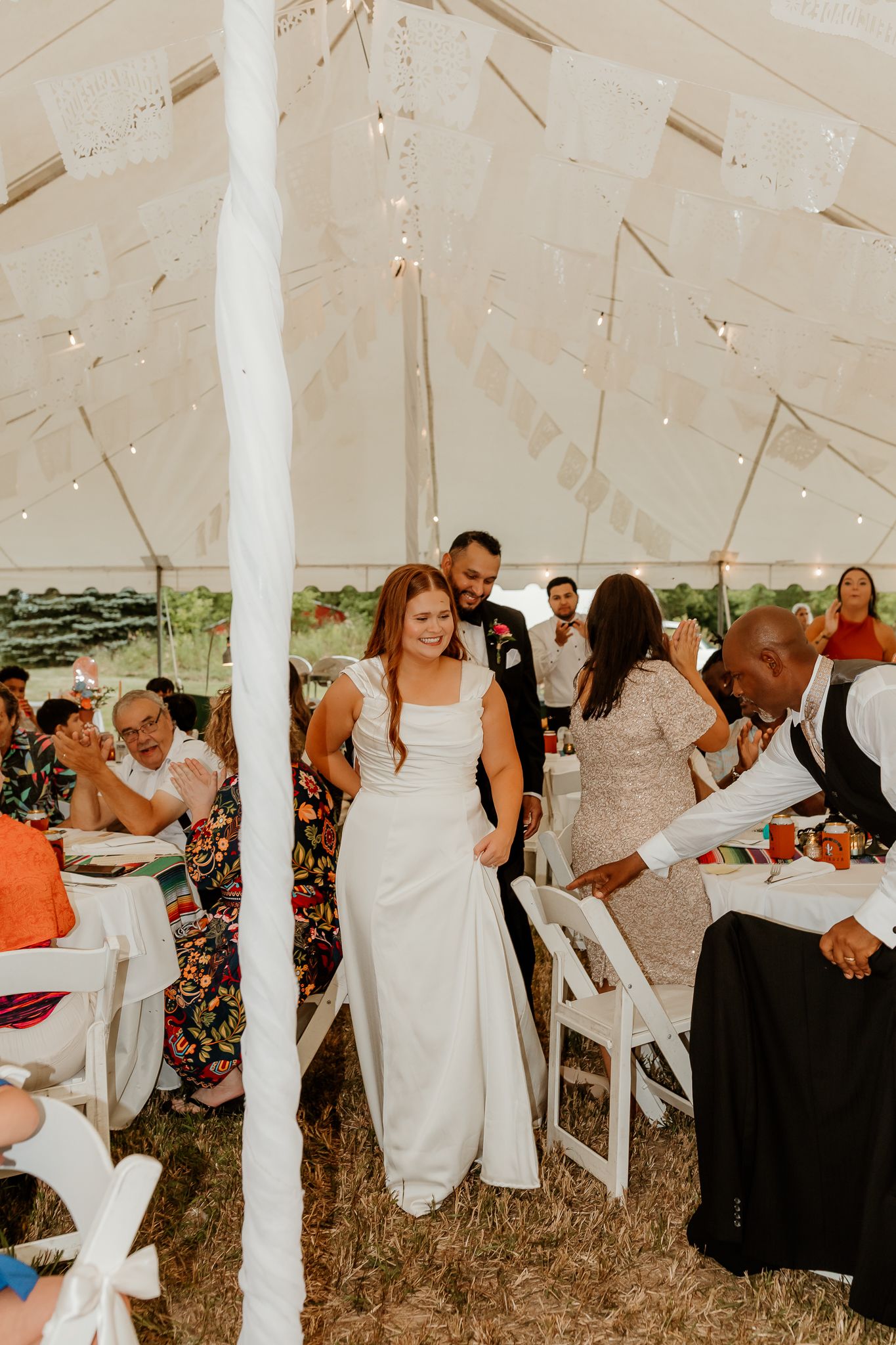 Bride in white gown walks through wedding reception tent with guests seated at decorated tables.