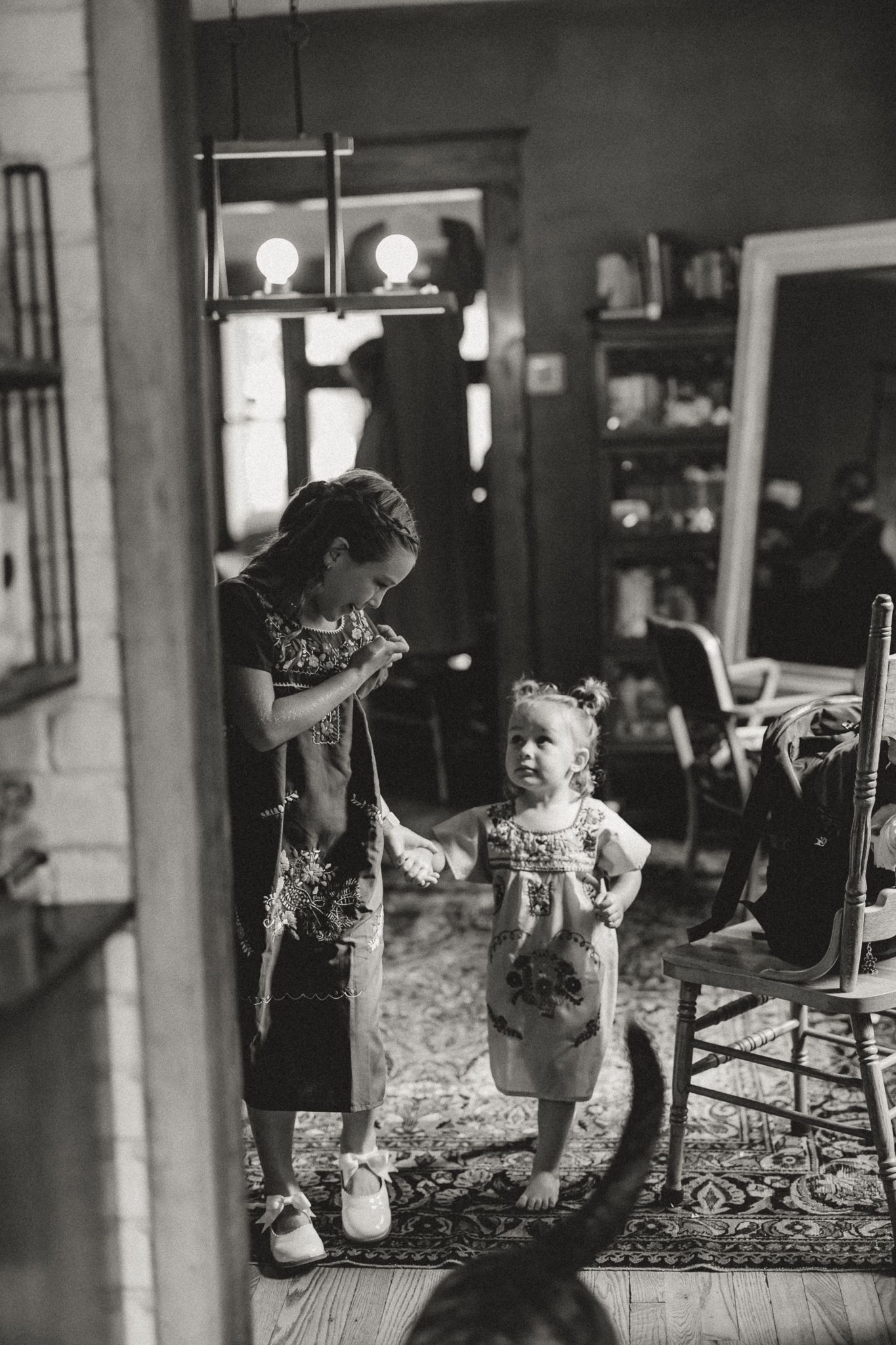 Black and white photo of an older girl helping dress a toddler in a vintage-style room with warm ambient lighting.