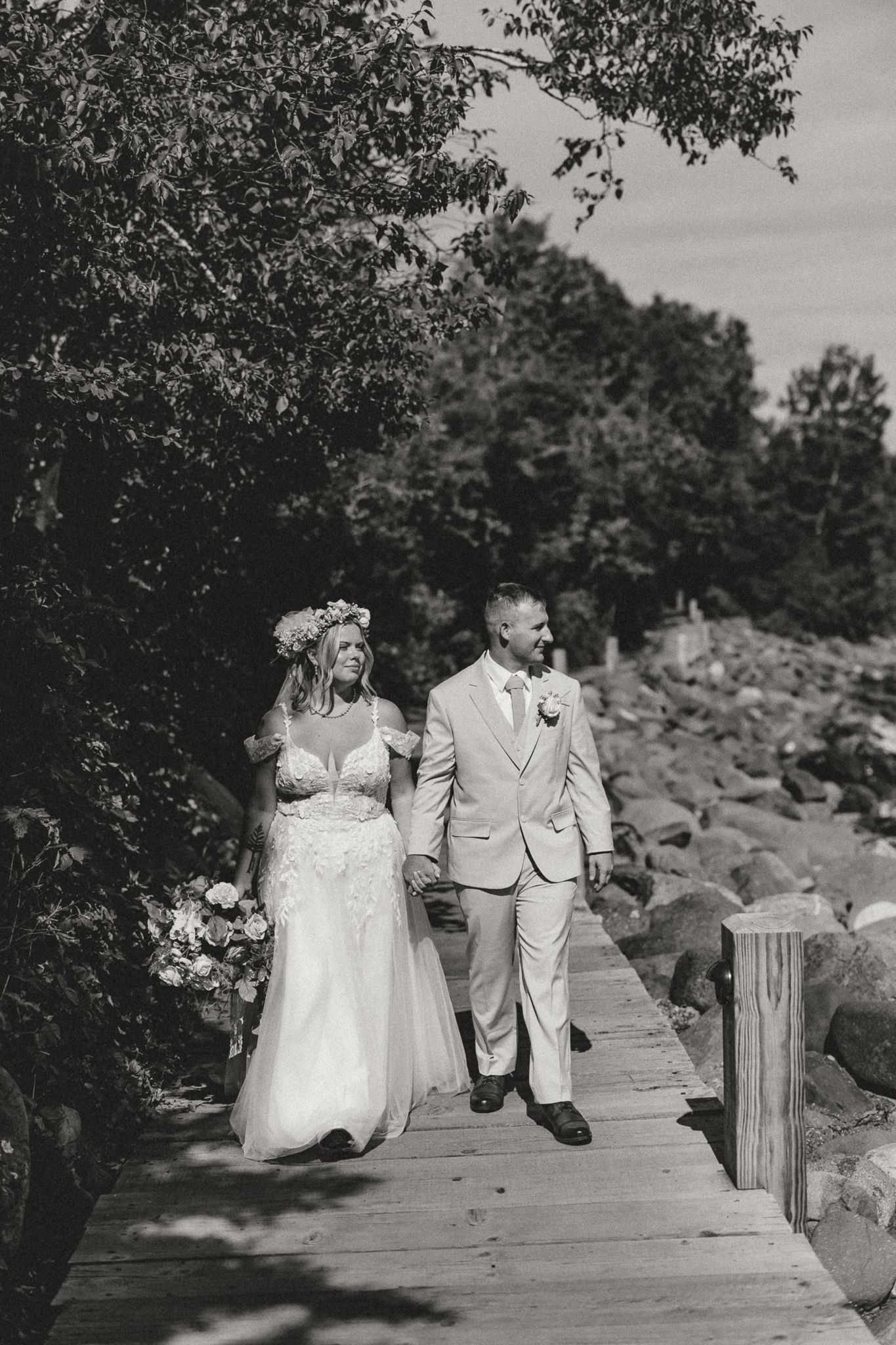 Black and white photo of a bride and groom walking hand in hand on a wooden dock surrounded by trees and rocks.
