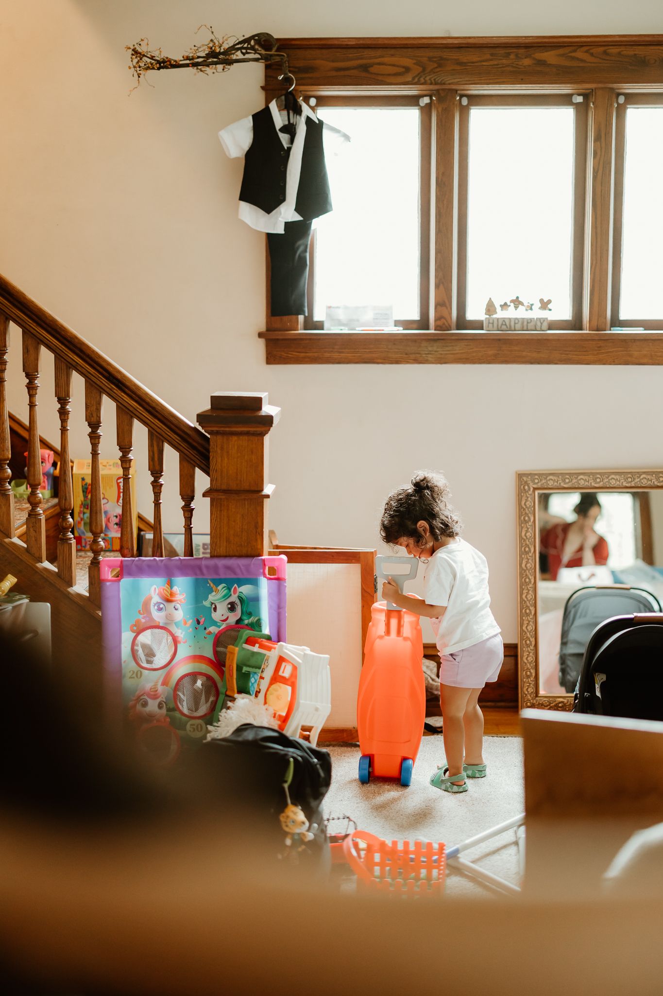A toddler plays with colorful toys near a staircase in a cozy home, with a small suit hanging above.