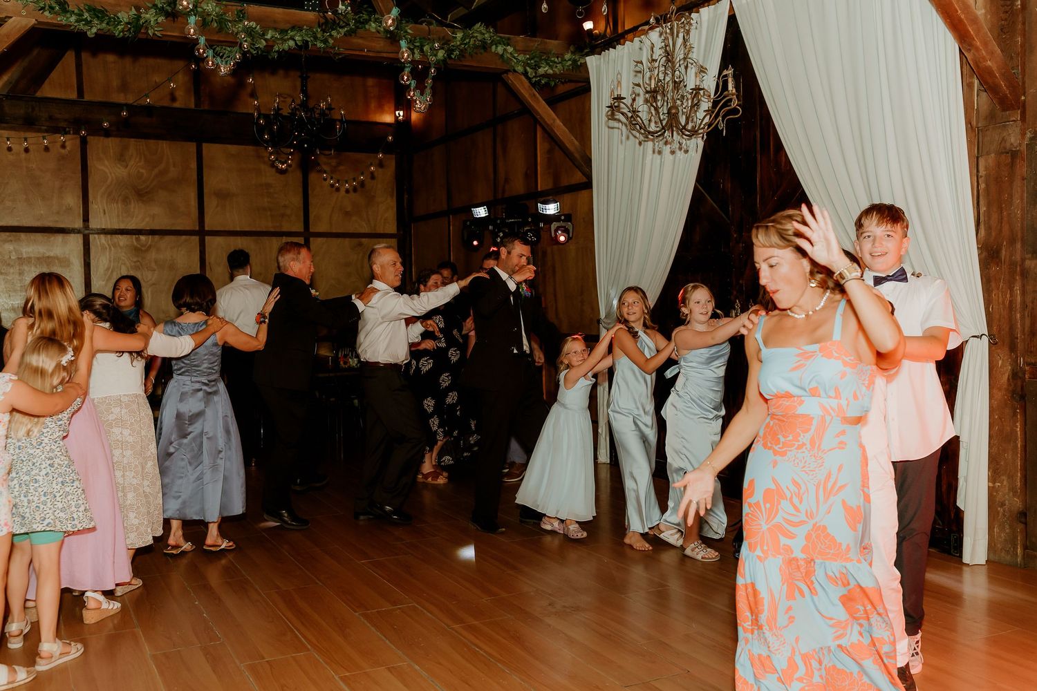 Guests dancing joyfully at a rustic barn wedding reception with chandelier lighting and elegant decorations.