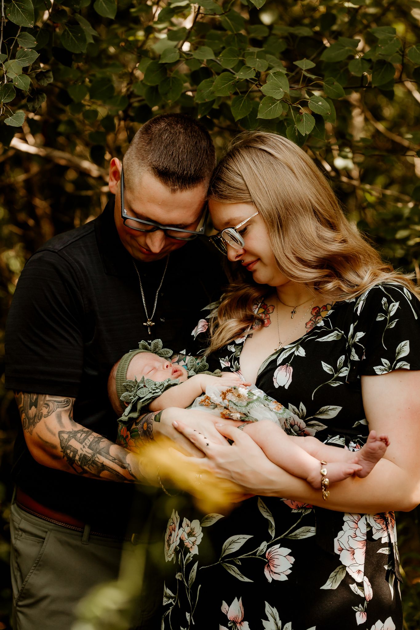A couple with tattoos and glasses tenderly cradles their newborn baby outdoors, surrounded by lush green foliage.