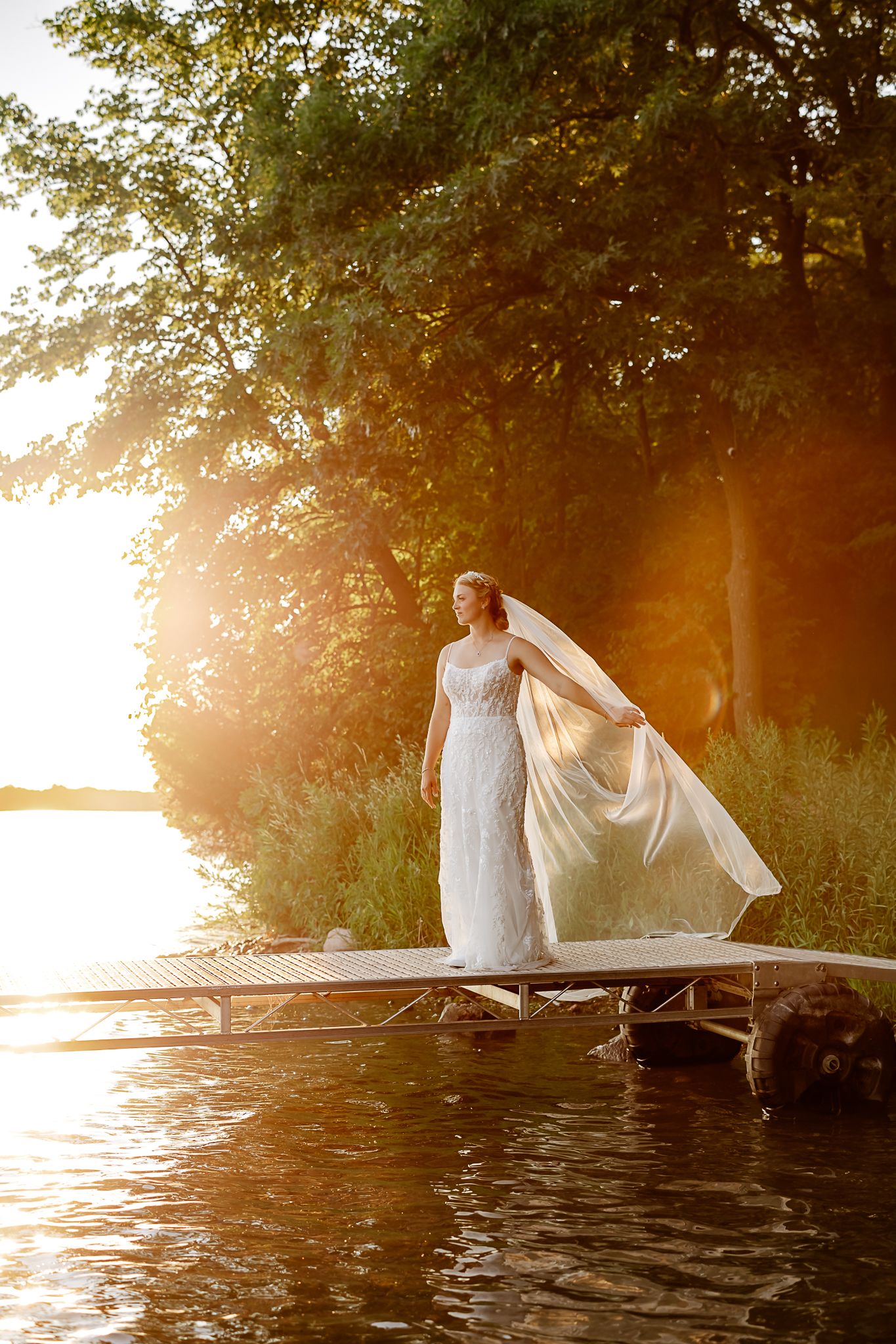 Bride in white gown and veil stands on lakeside dock at golden sunset surrounded by lush green trees.