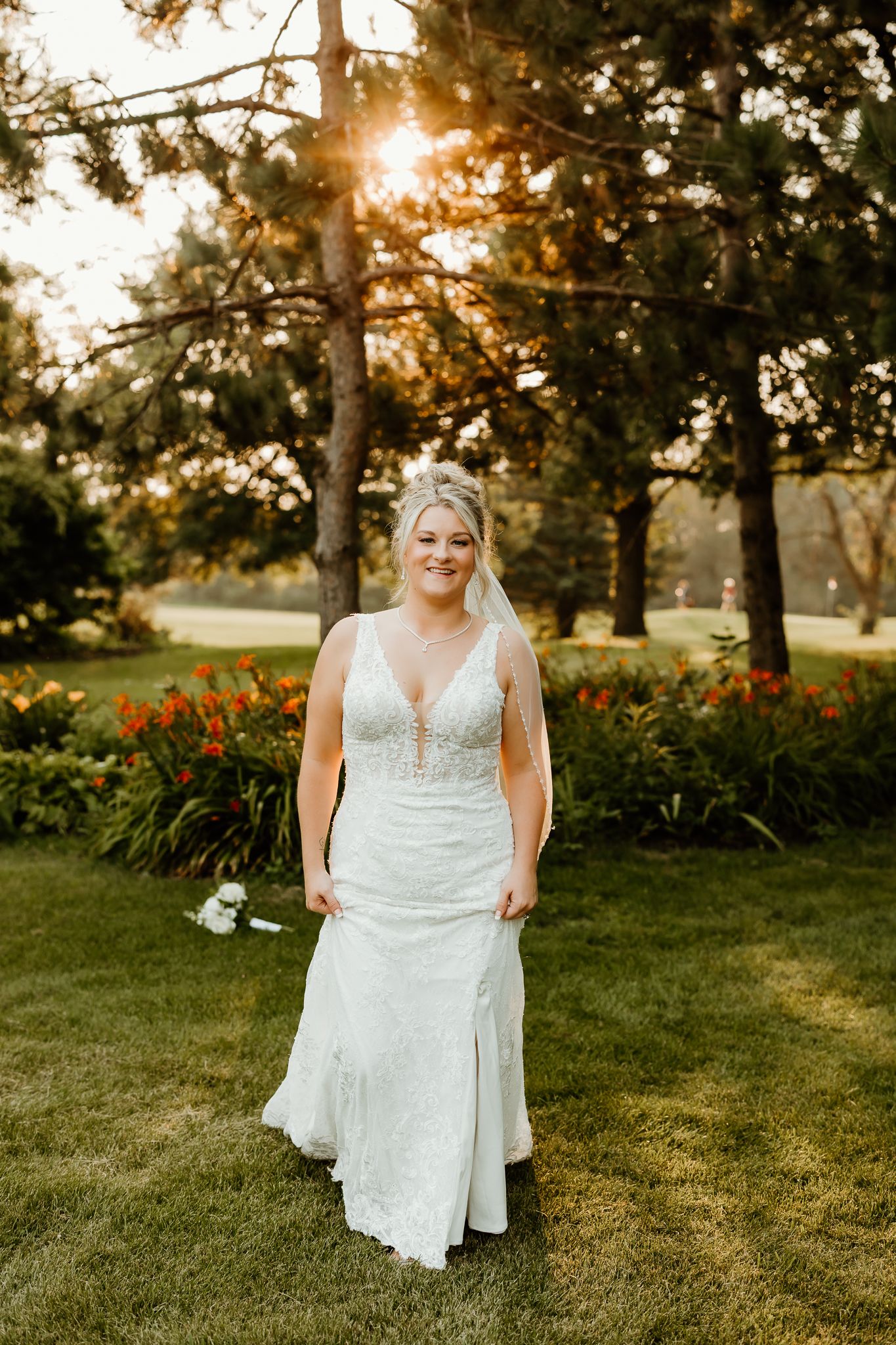 Bride in white lace gown stands on green lawn at golden hour with colorful flowers and trees in background.