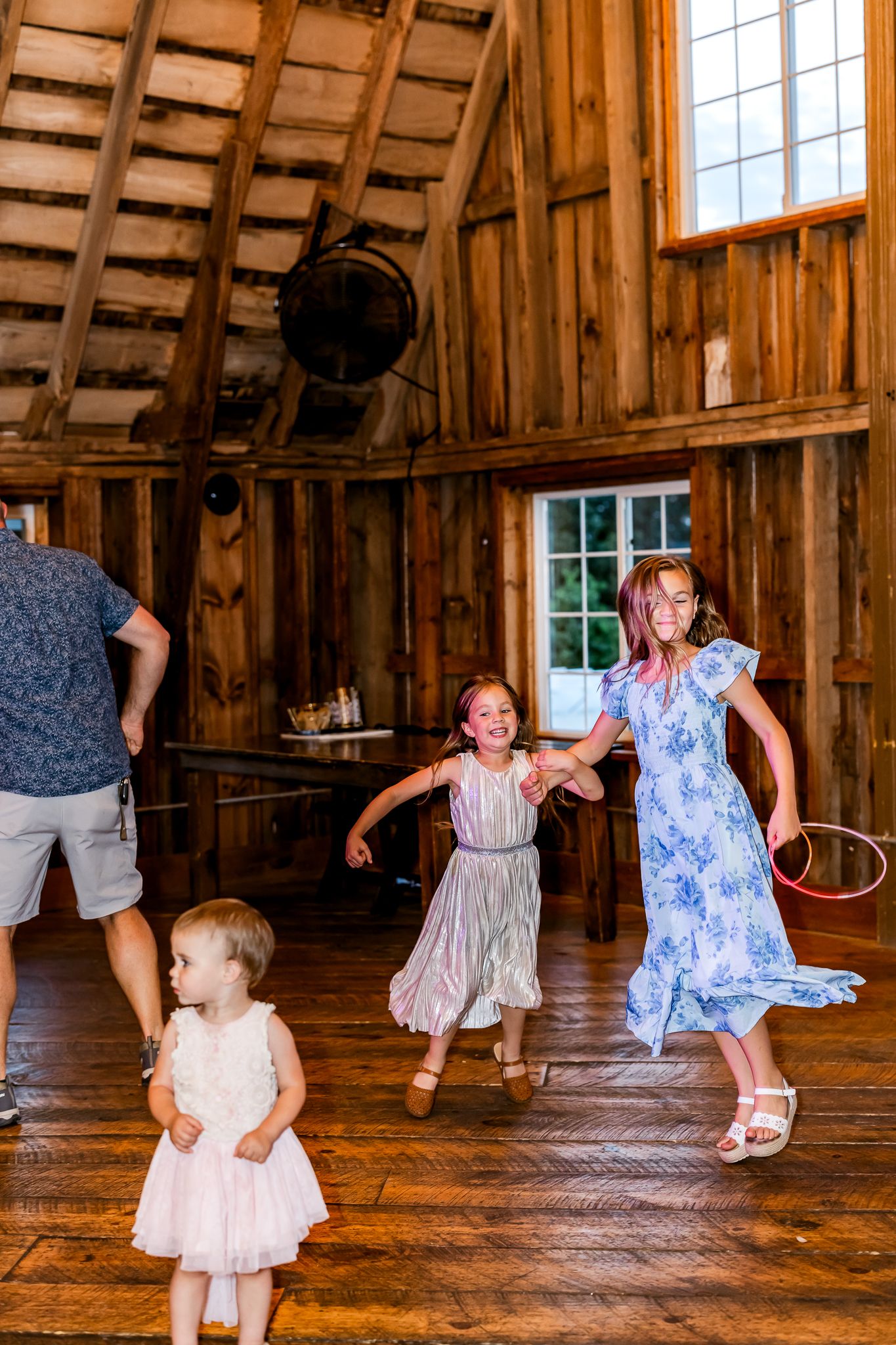 Children dancing joyfully in a rustic wooden barn venue during a celebration, wearing colorful dresses on hardwood floors.