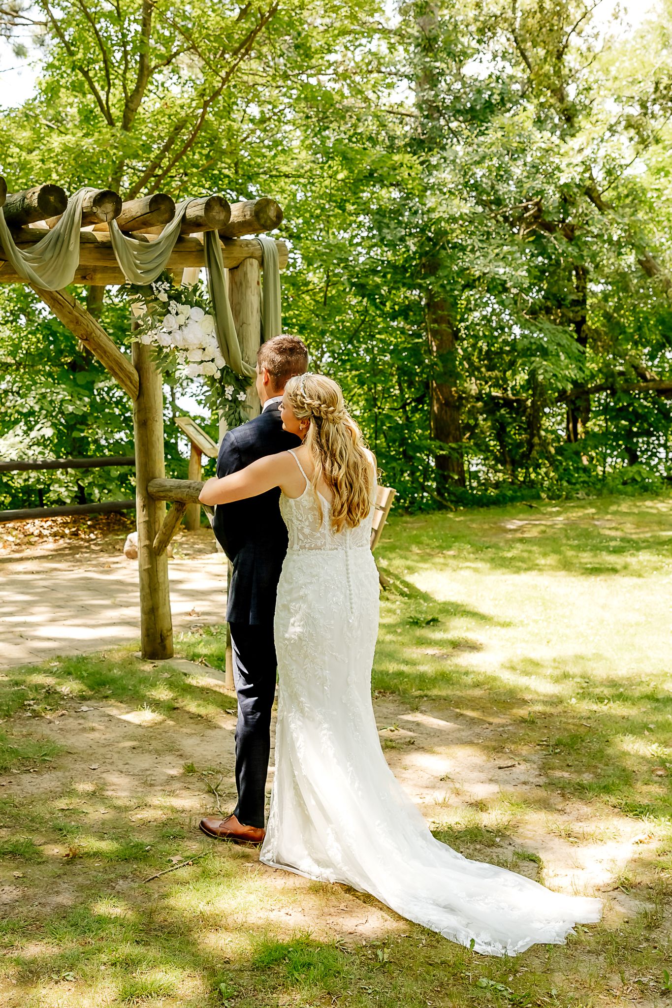 Bride and groom standing together at outdoor rustic wooden wedding arch surrounded by lush green trees.
