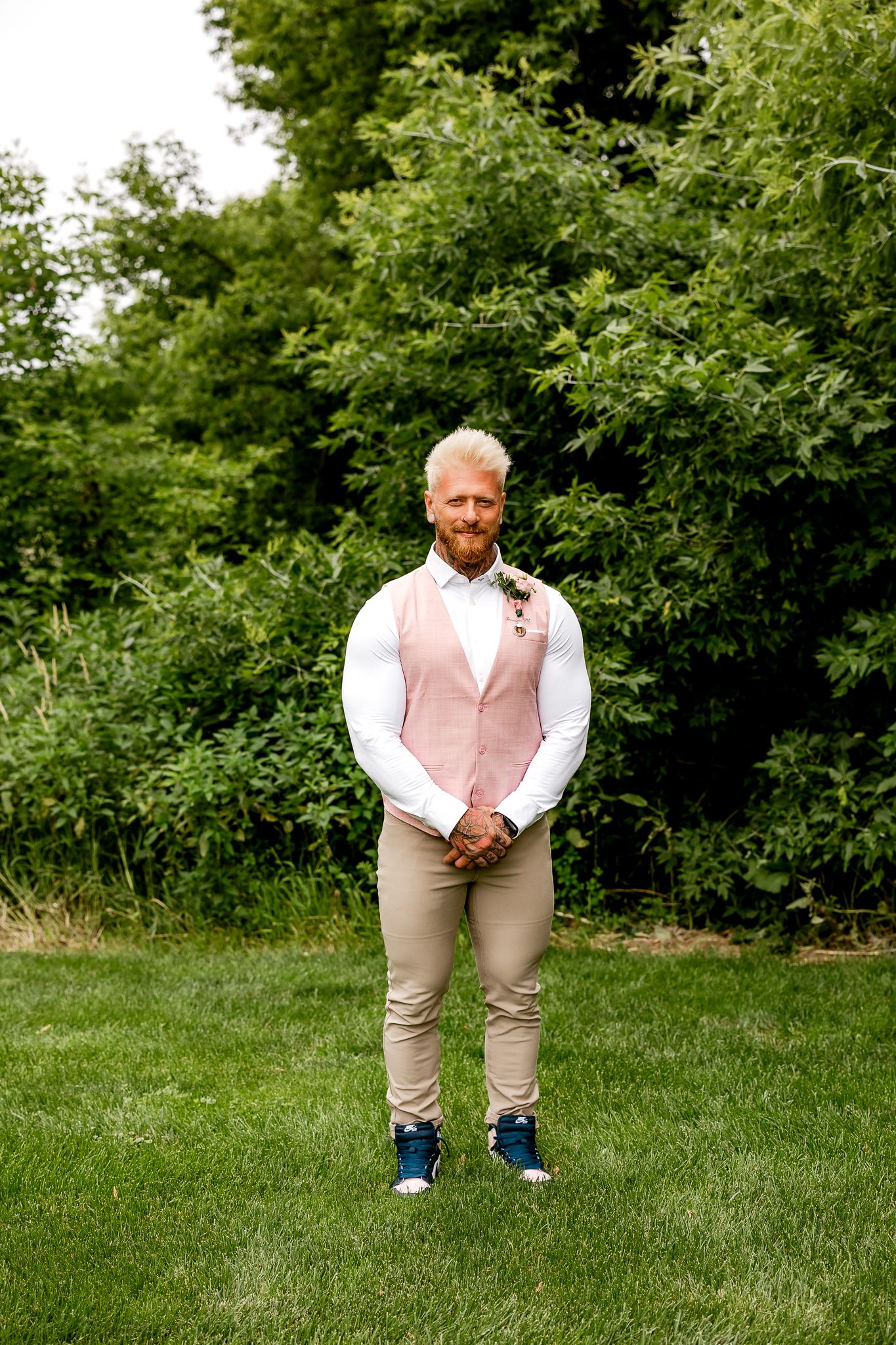 A smiling groom in a pink vest, white shirt, and tan pants stands on green grass with lush trees behind him.