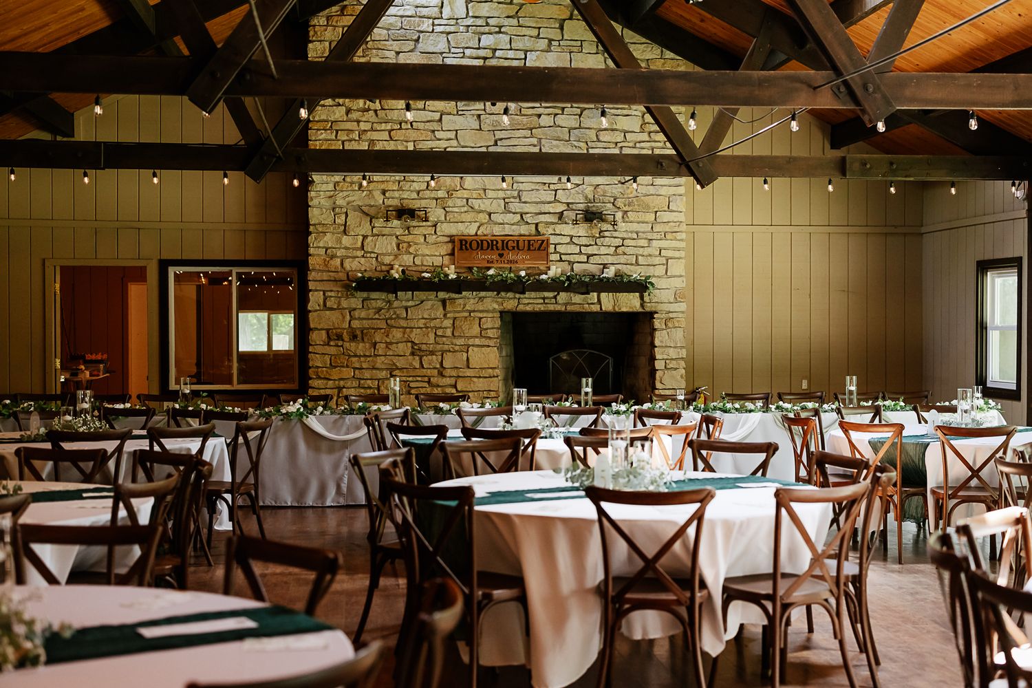 Rustic wedding reception hall with stone fireplace, round tables, white linens, and wooden cross-back chairs under string lights.