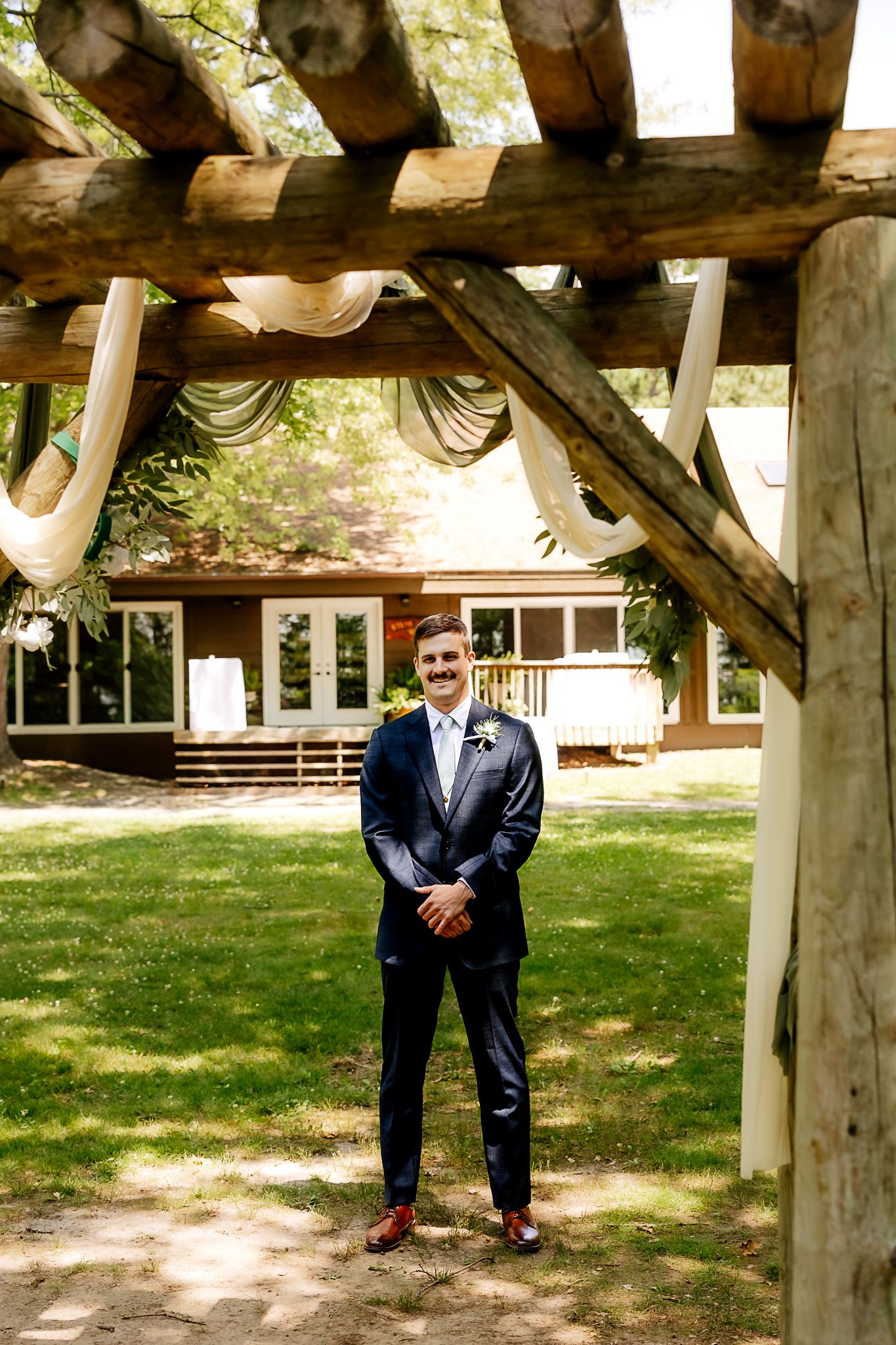 Groom in navy suit standing under rustic wooden pergola with draped fabric at outdoor wedding venue.