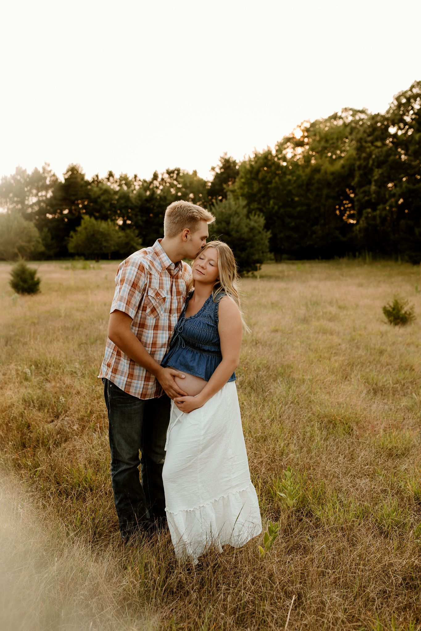 A couple shares a tender kiss in a sunlit field, the woman in a white dress cradling her baby bump.