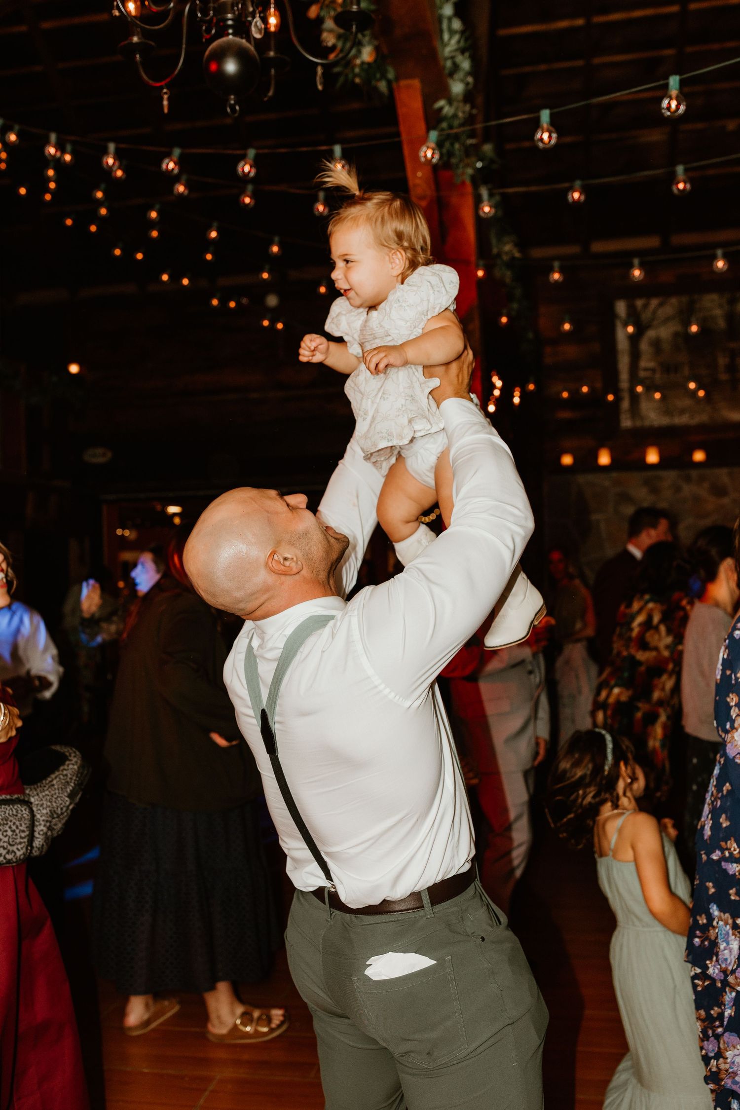 Bald man in white shirt and suspenders lifts a laughing toddler in a white dress overhead on a dance floor under string lights.