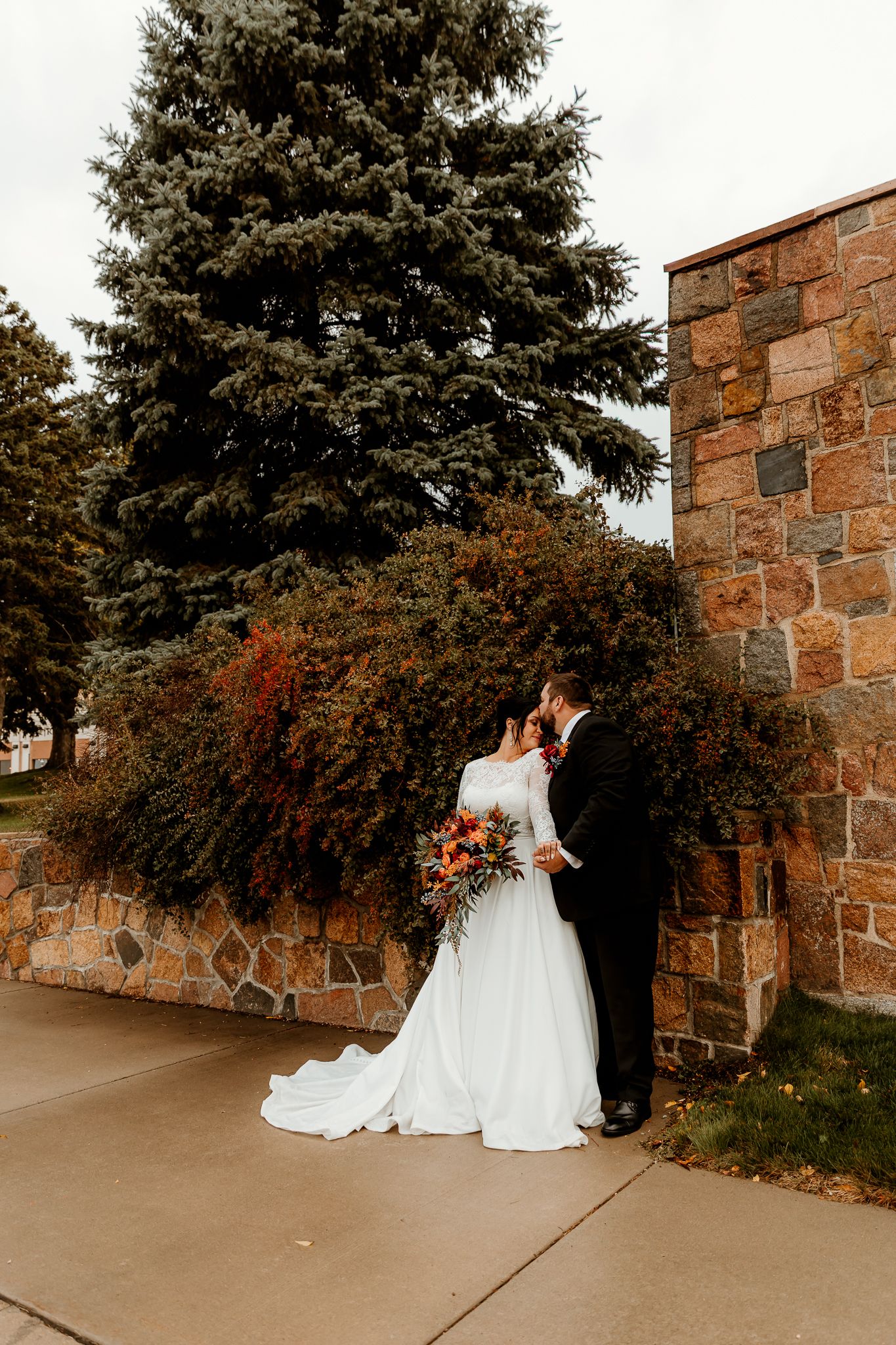 Bride in a long white gown holding an autumn bouquet embraces her groom in a black suit beside a stone wall and evergreen tree.