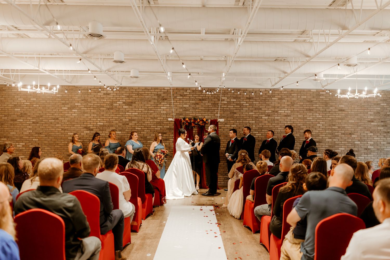 Bride and groom exchange wedding vows at the altar before seated guests in red chairs along a white aisle runner in a brick-walled venue.