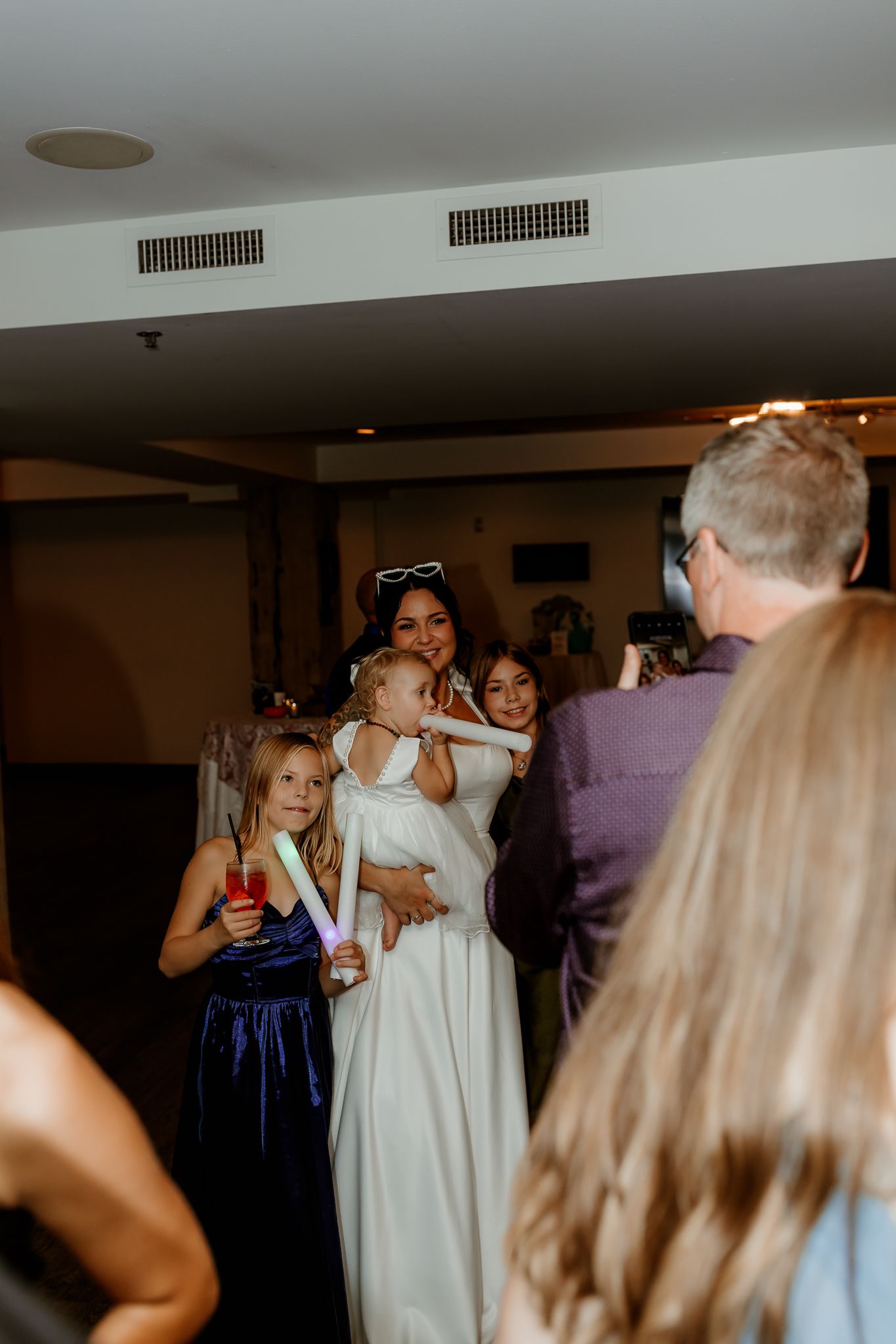 Bride in a white gown holding a toddler while a girl in a navy dress holds glow sticks at a lively indoor wedding reception.