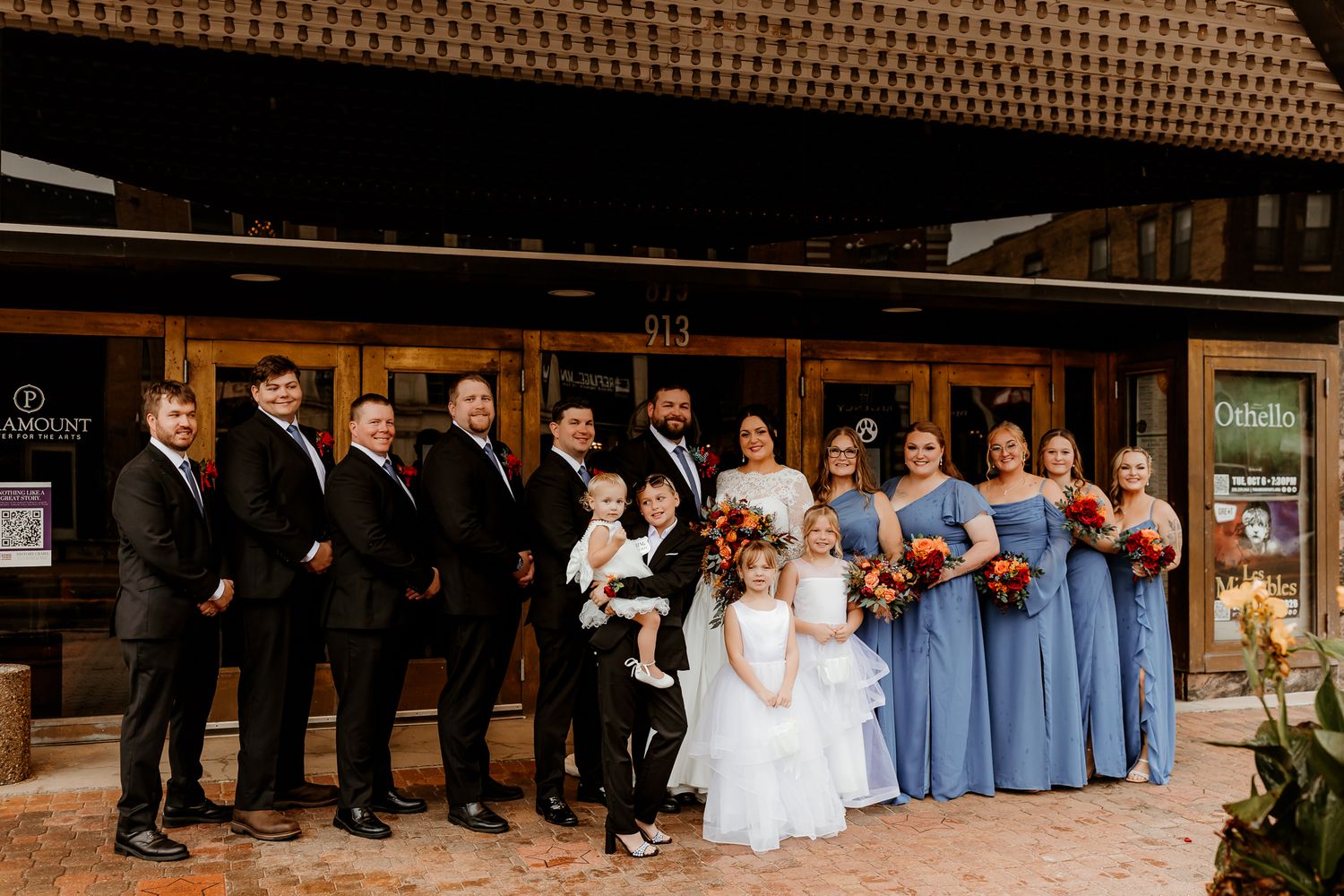 Wedding party in black suits and dusty blue bridesmaid dresses poses with a bride, flower girl and ring bearer outside a brick building.