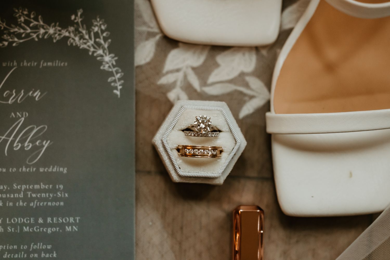 Wedding flat lay with stacked engagement and wedding rings in a hexagonal ring box beside white heels and a sage green invitation.