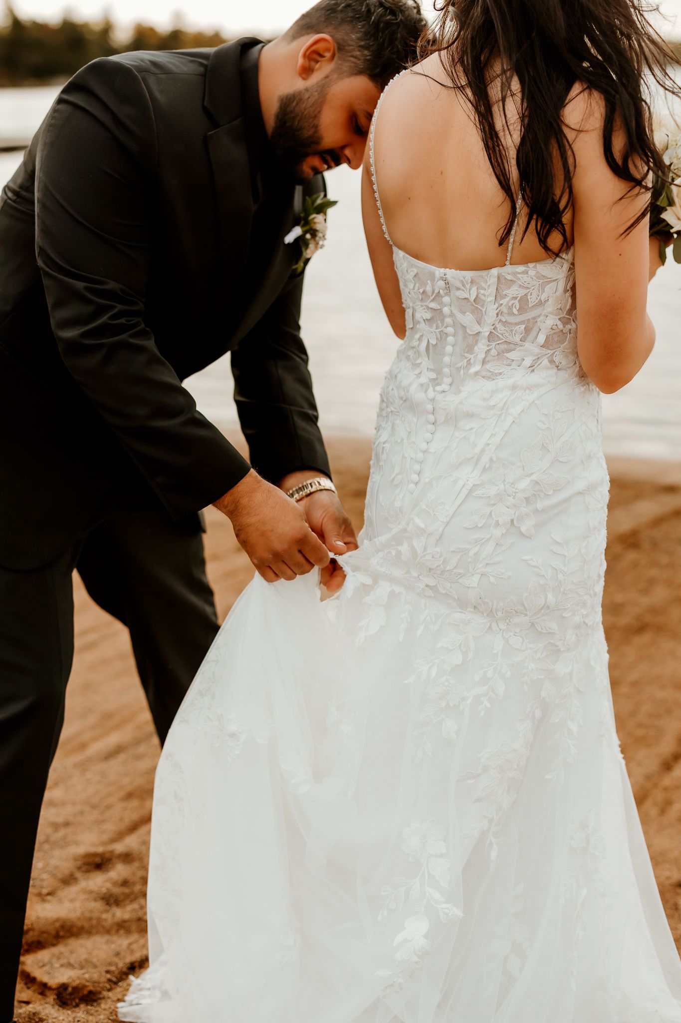 Groom in a black suit bends down to adjust the lace skirt of a bride's white wedding gown on a sandy lakeside beach.
