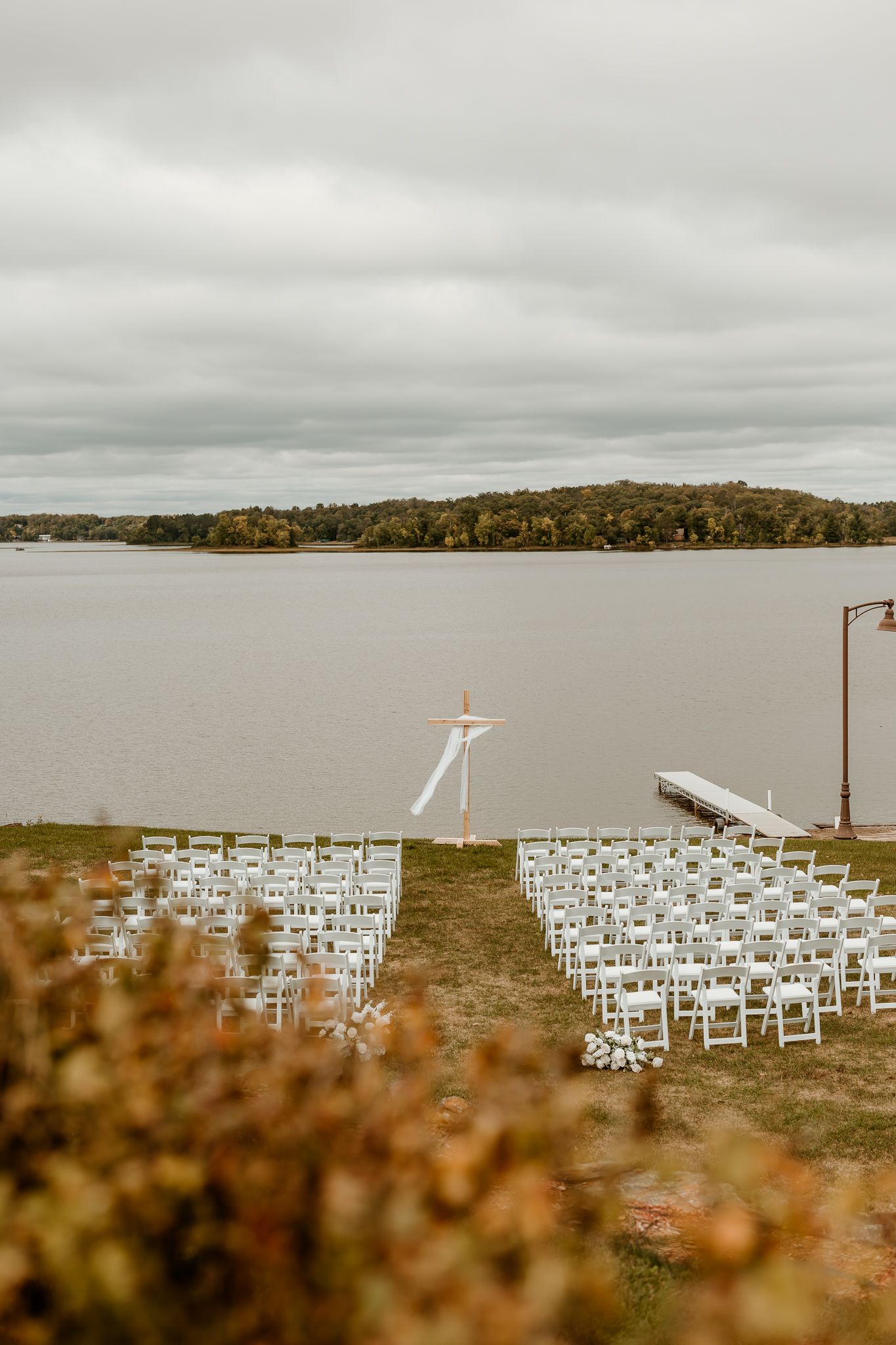 Outdoor lakeside wedding ceremony setup with rows of white chairs, a wooden arch, and a dock under an overcast autumn sky.