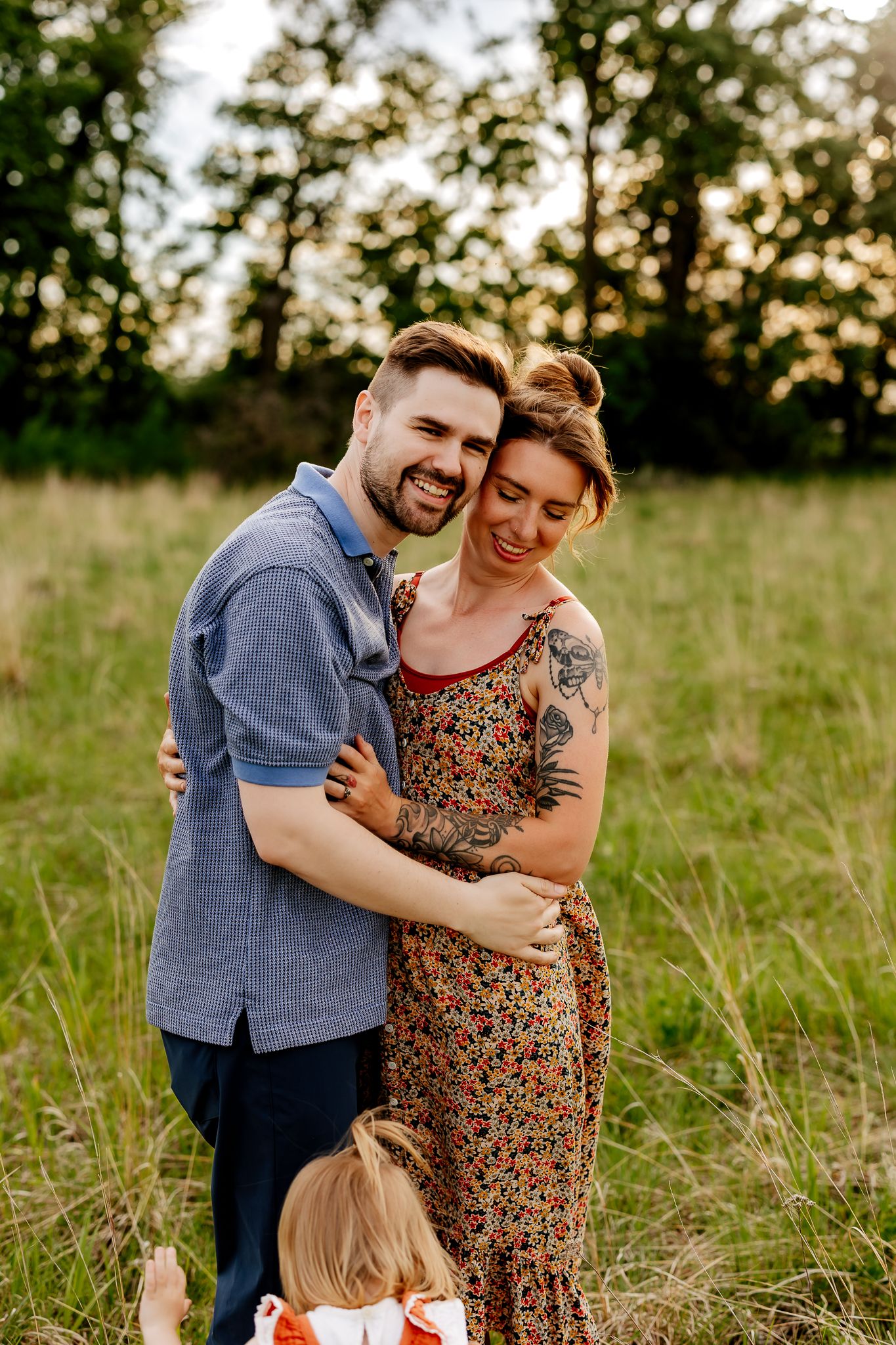 A smiling couple embraces in a golden-lit meadow while a young child stands in front of them.