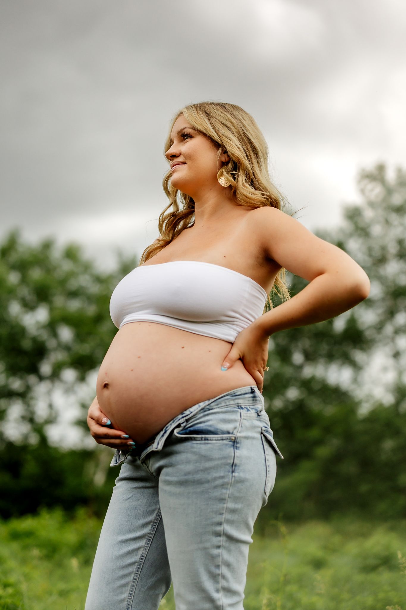 Smiling pregnant blonde woman cradling baby bump in white tube top and jeans in nature setting.