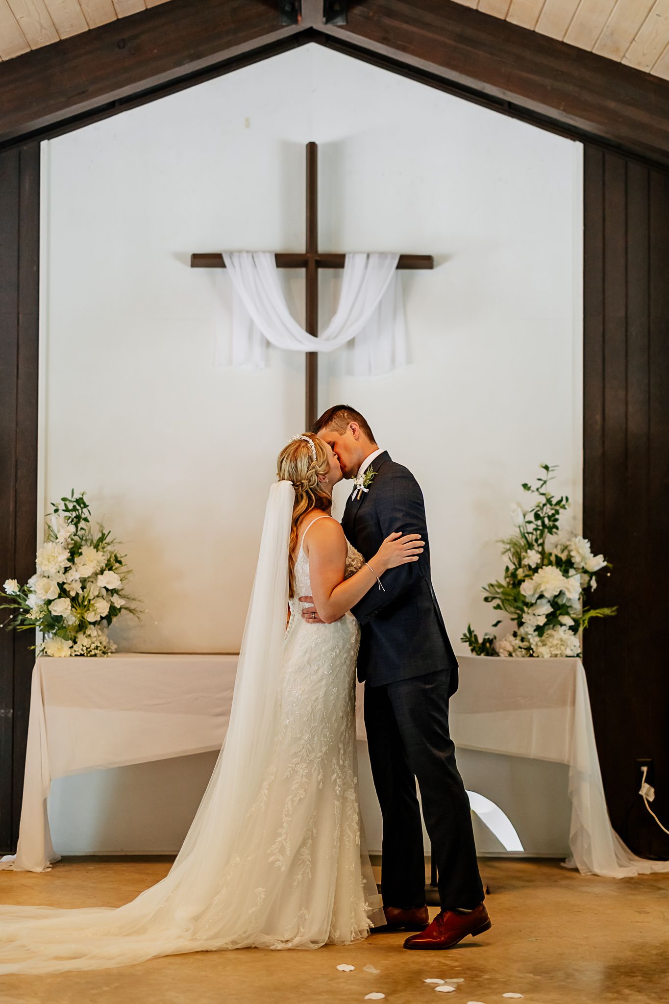 Bride and groom share first kiss at chapel altar with wooden cross, white draping, and floral arrangements.