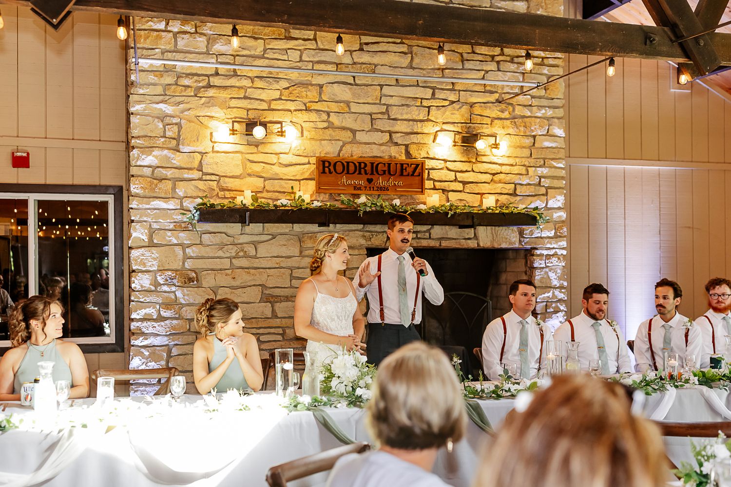 Bride and groom share a toast with wedding party near stone fireplace in rustic reception venue.