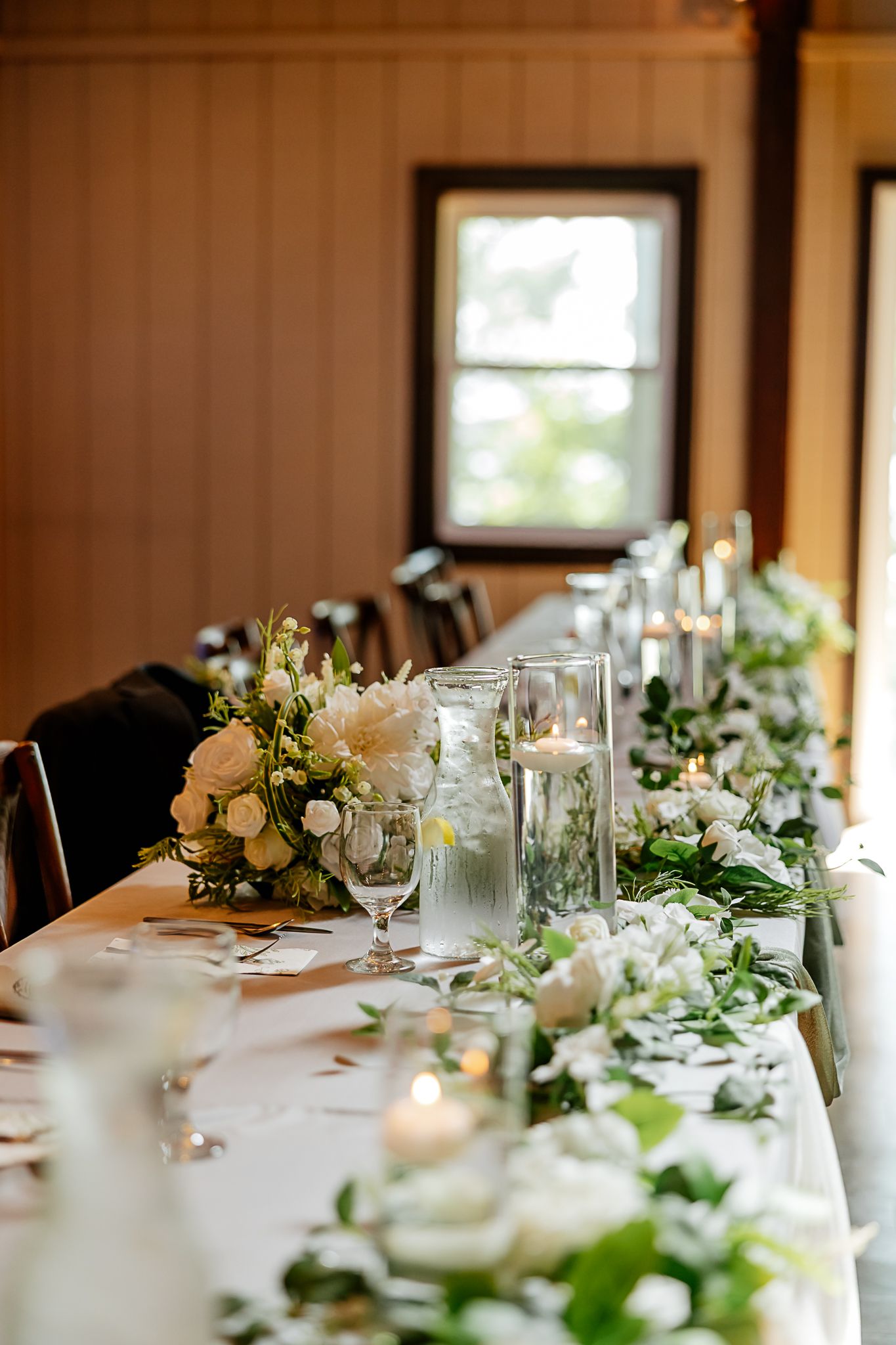 Elegant wedding reception table with white floral centerpieces, greenery runner, and glowing candles in a rustic wood-paneled venue.