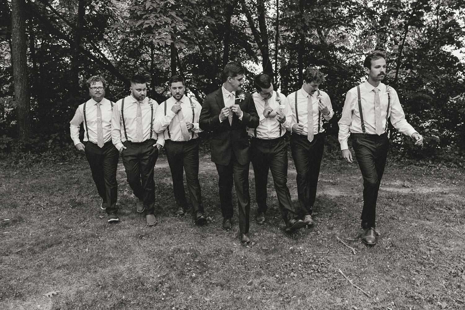 Black and white photo of a groom and groomsmen in suspenders walking outdoors near trees.