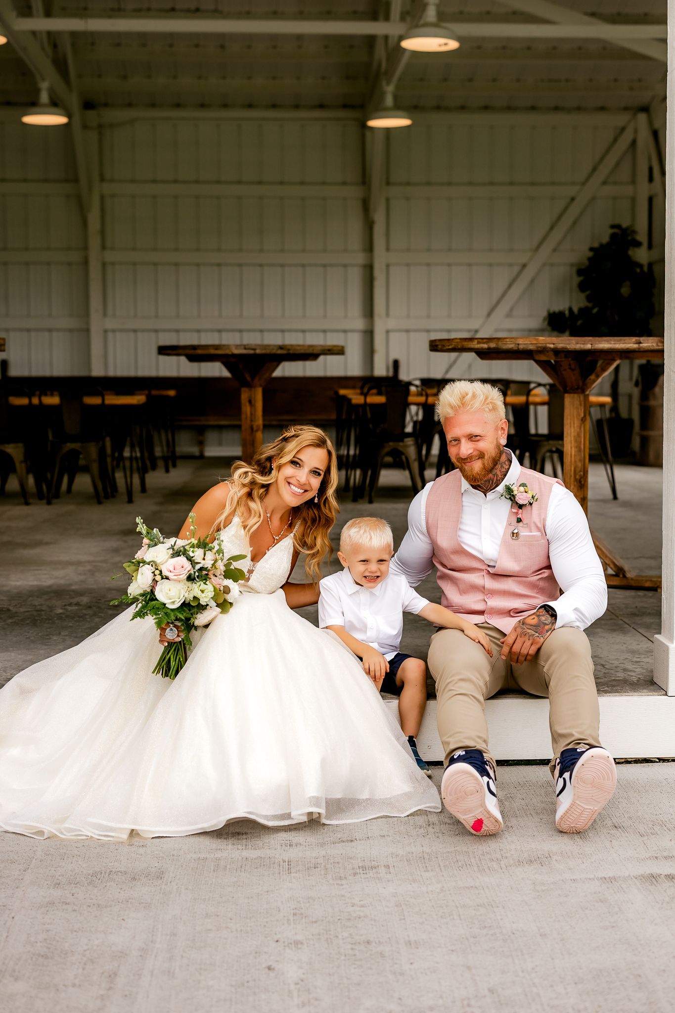 Bride in white gown and groom in pink vest sit with young boy in barn wedding venue holding floral bouquet.