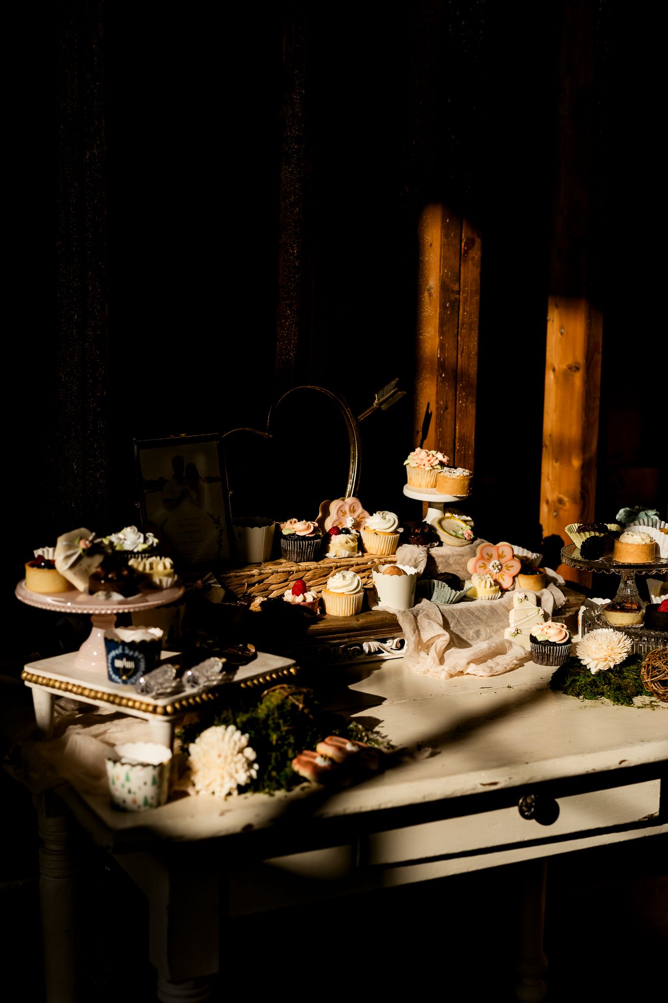 Rustic dessert table with cupcakes, macarons, and pastries displayed on vintage stands in moody, dramatic lighting.