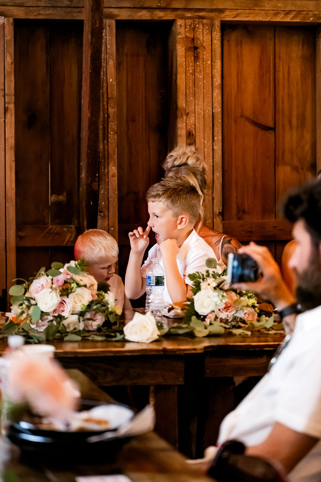 Two young boys sit at a rustic wooden table adorned with floral centerpieces at a wedding reception.