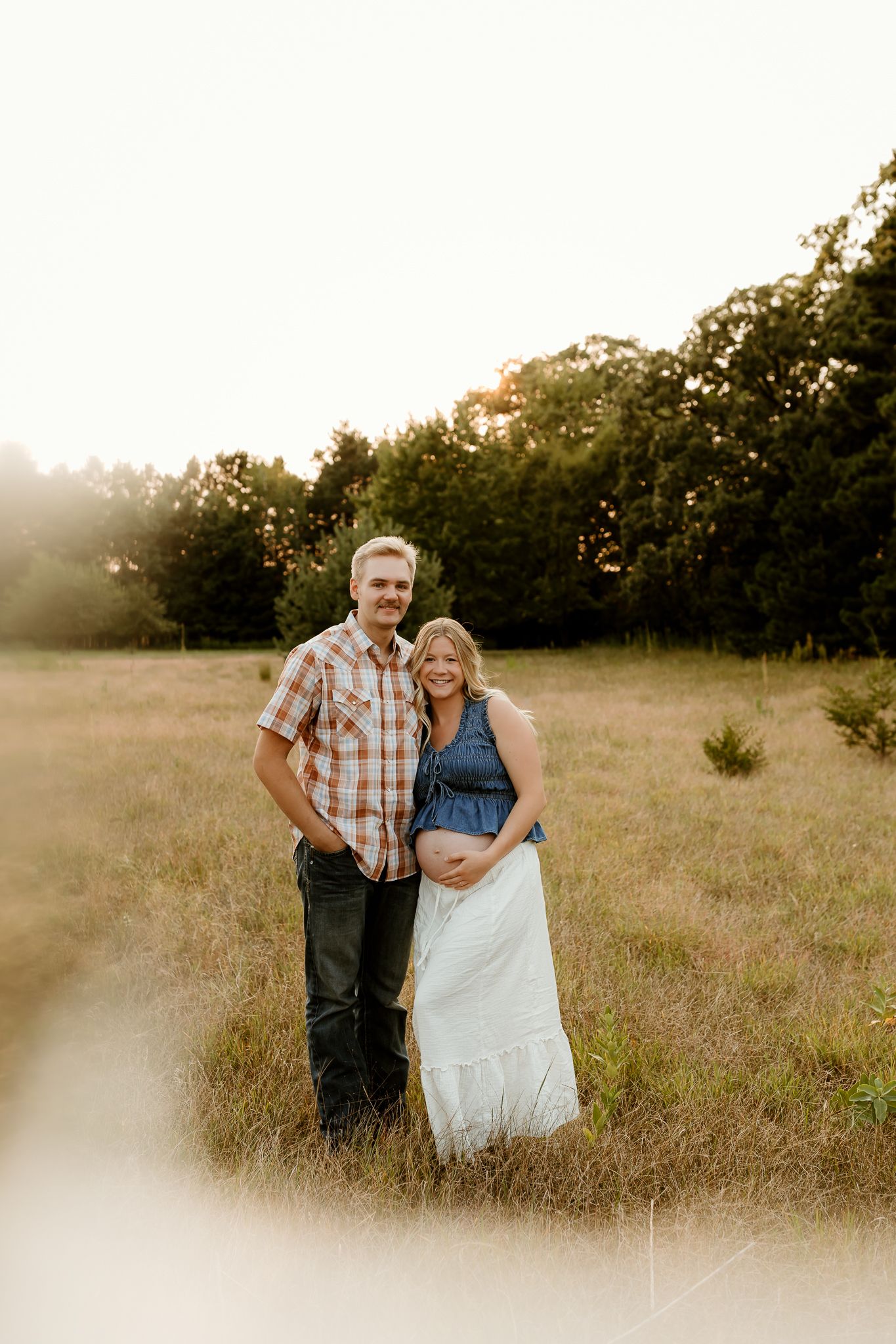 Couple poses in golden field at sunset, pregnant woman in denim top and white skirt, man in plaid shirt.