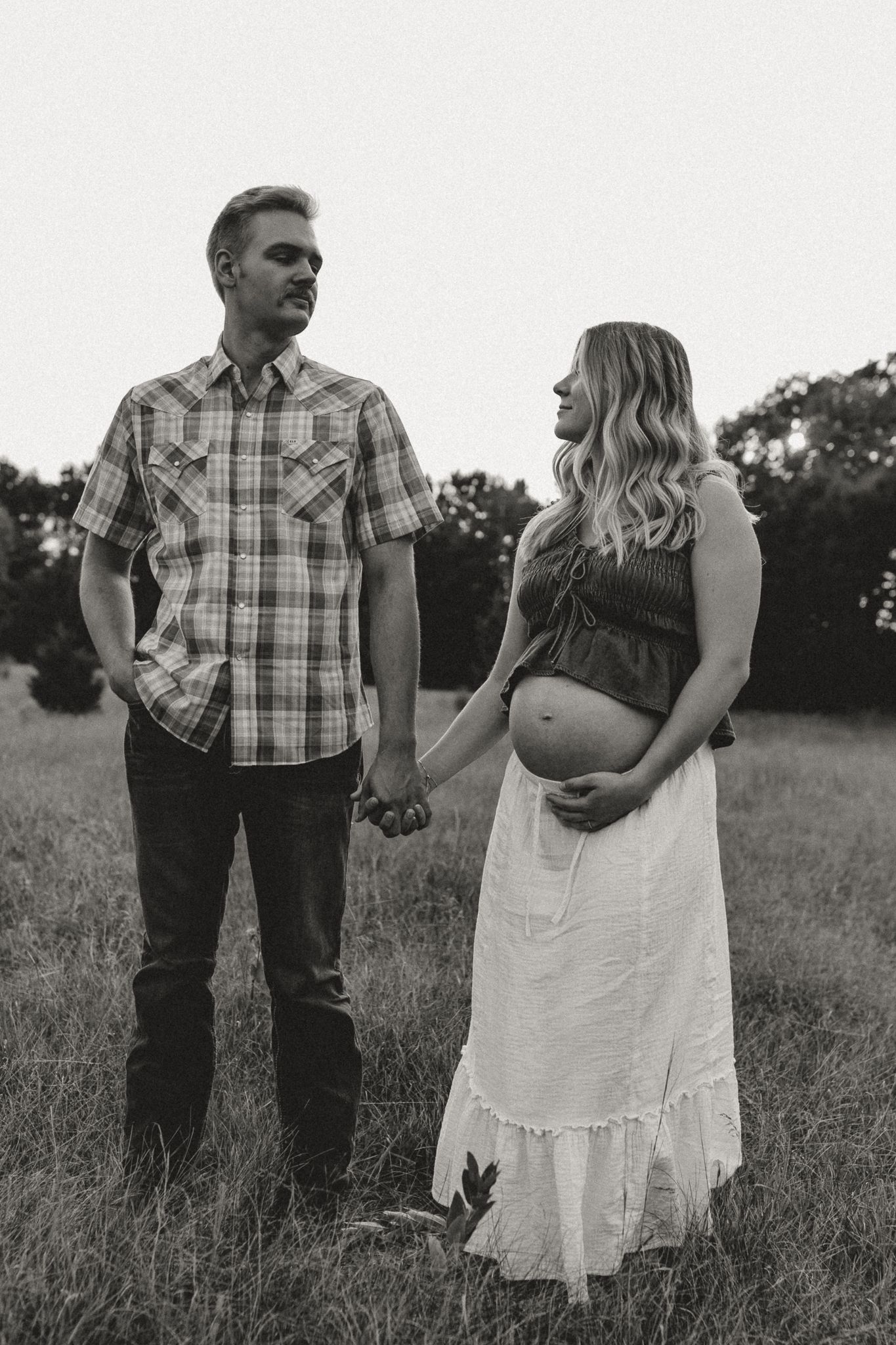 Black and white maternity photo of a couple holding hands in a field, woman cradling baby bump in white skirt.