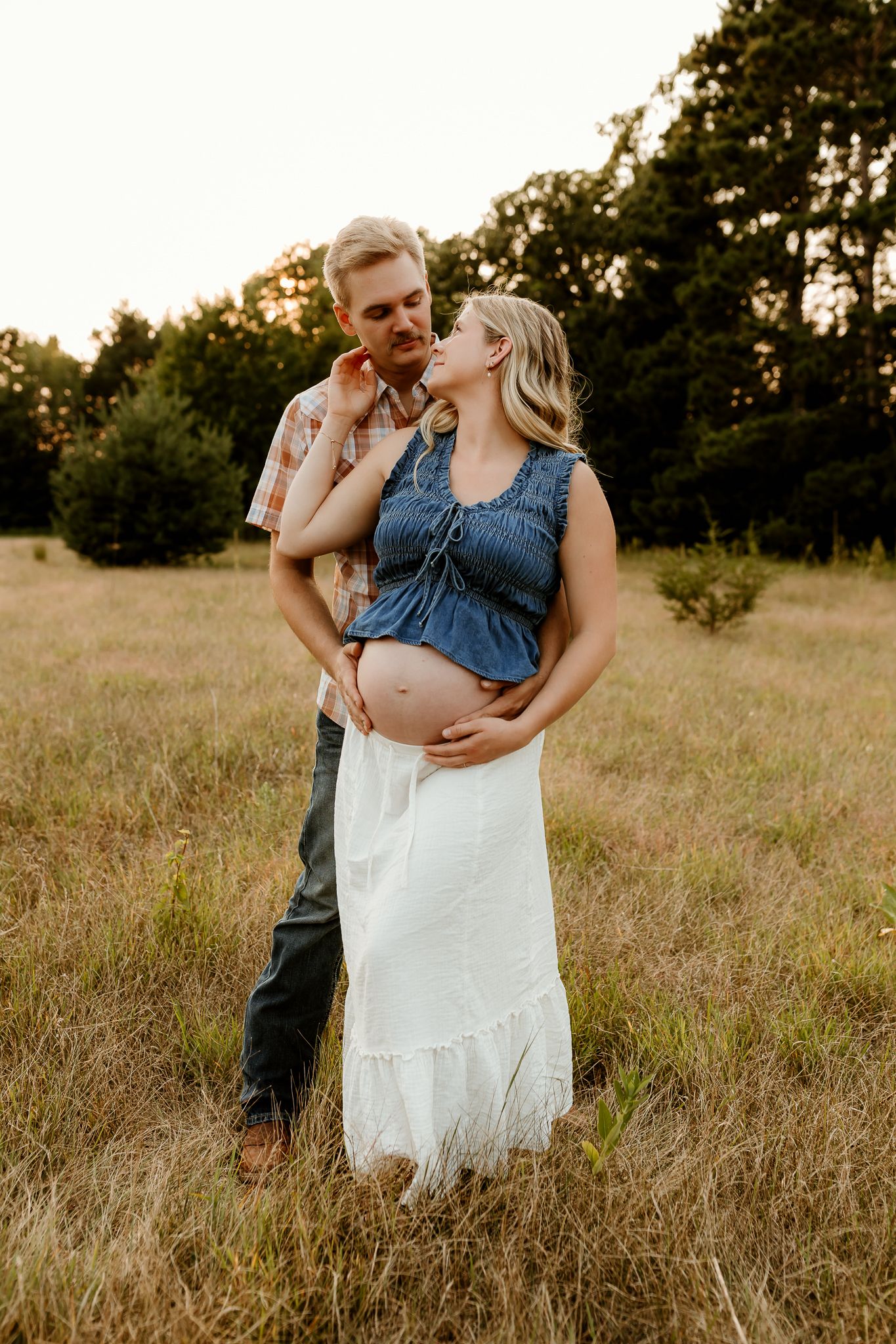 Couple sharing a tender moment in a field at golden hour, woman in denim top and white skirt cradling baby bump.