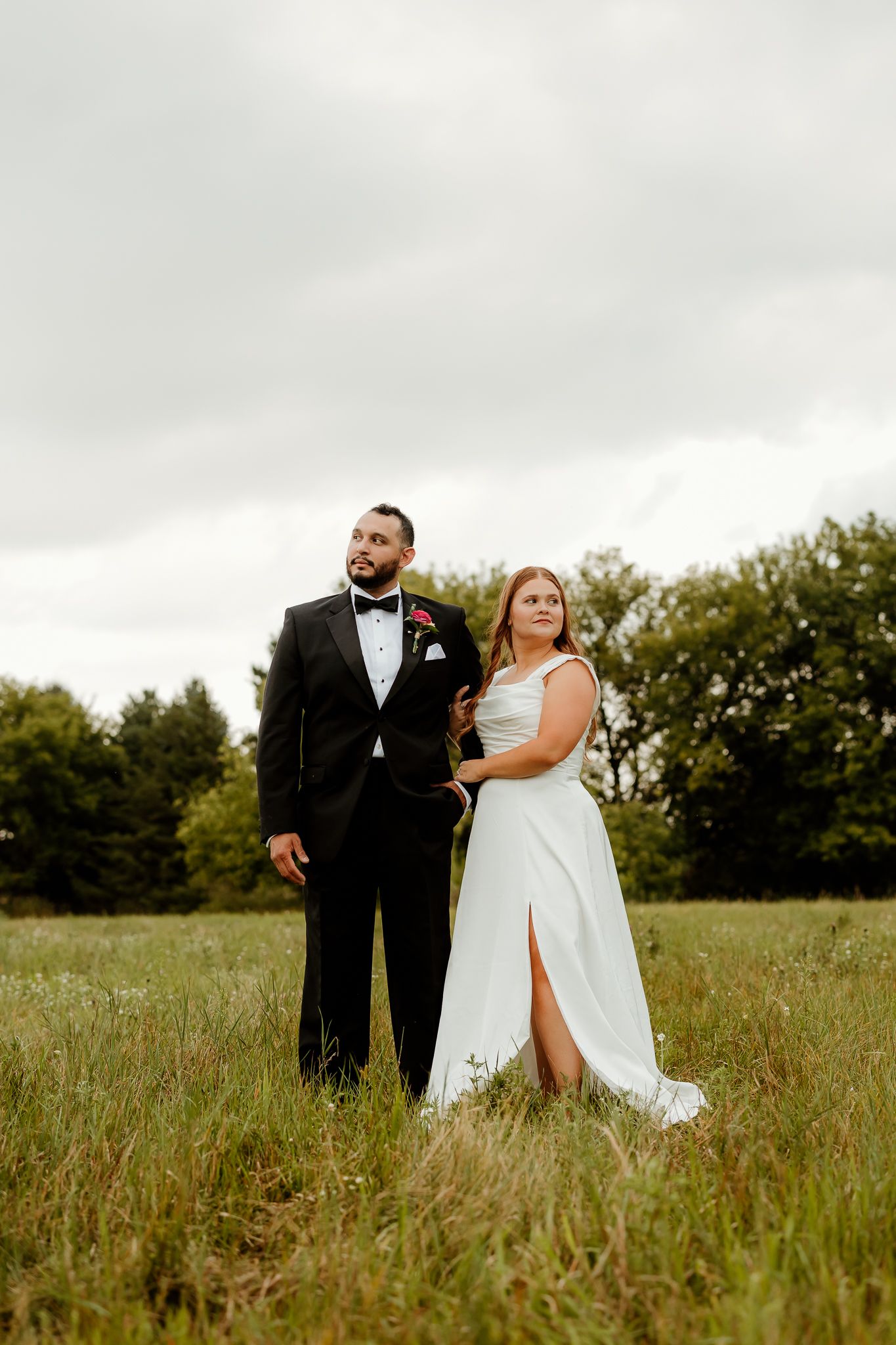 Bride in white gown and groom in black tuxedo standing together in a lush green field on their wedding day.