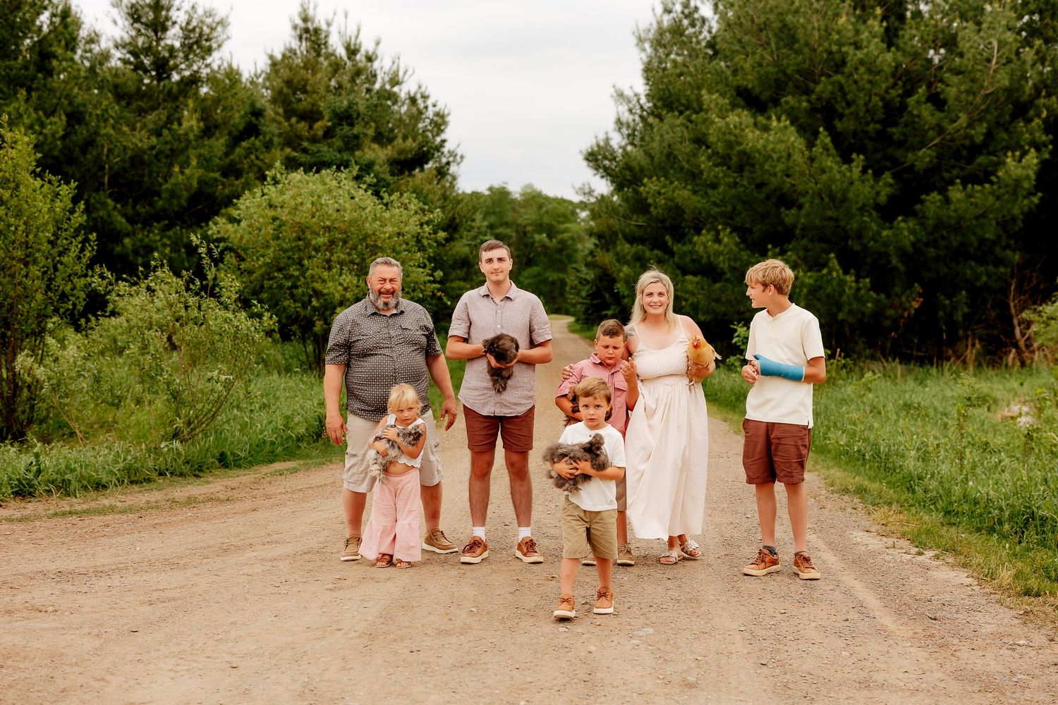 A large extended family of seven walks together down a dirt path surrounded by lush green trees.