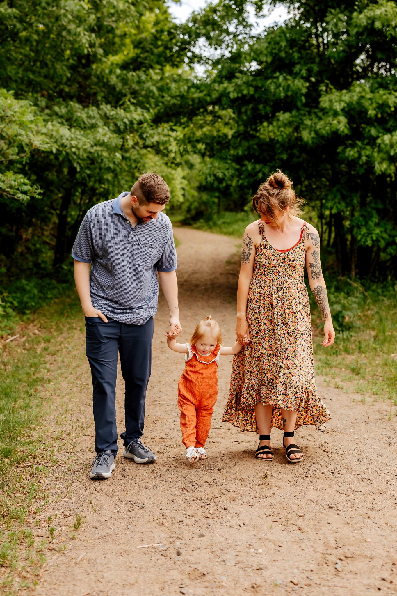 A family of three walks together on a nature trail, parents holding their toddler's hands in orange overalls.