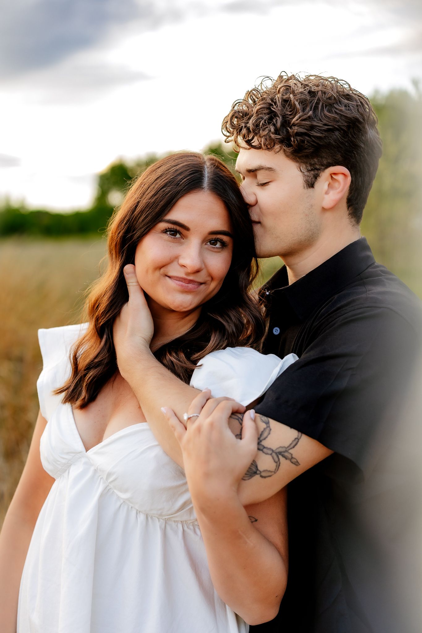 A young man tenderly kisses a smiling womans cheek while embracing her in an open field at sunset.
