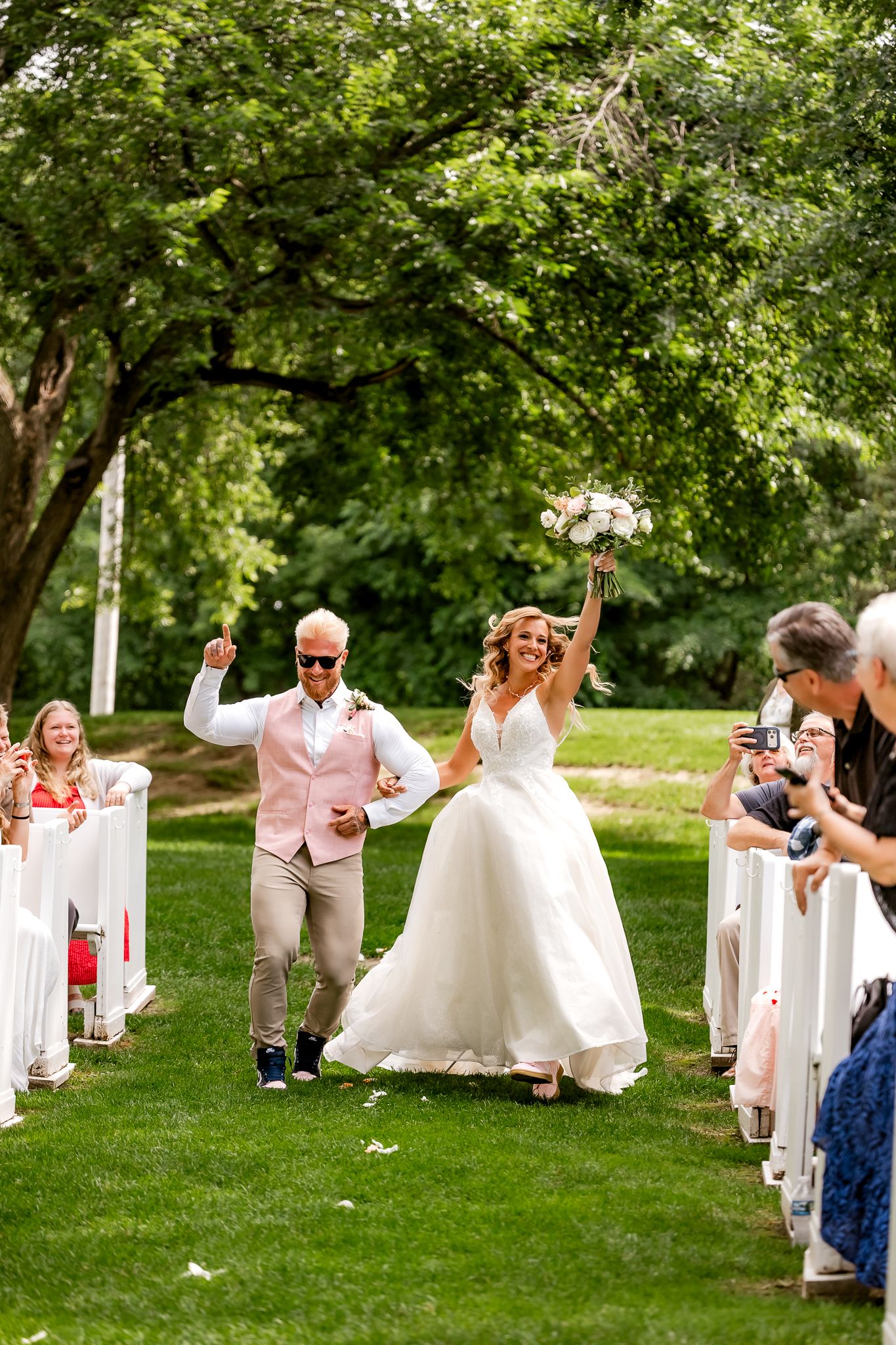 Joyful bride lifting bouquet and groom cheering as they walk back down the aisle after outdoor wedding ceremony.